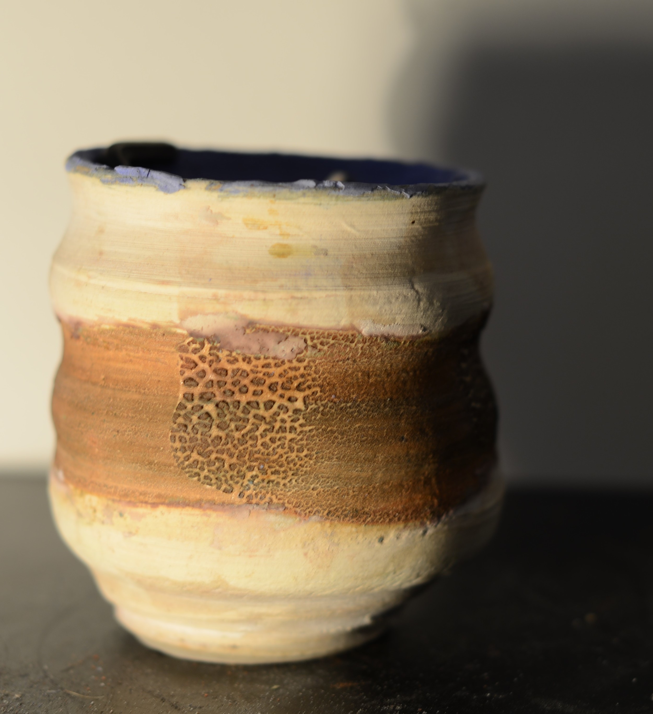 A ceramic pot with a textured, wavy surface and a blend of earthy colors including beige, orange, and brown, illuminated by sunlight with a shadow on the right.