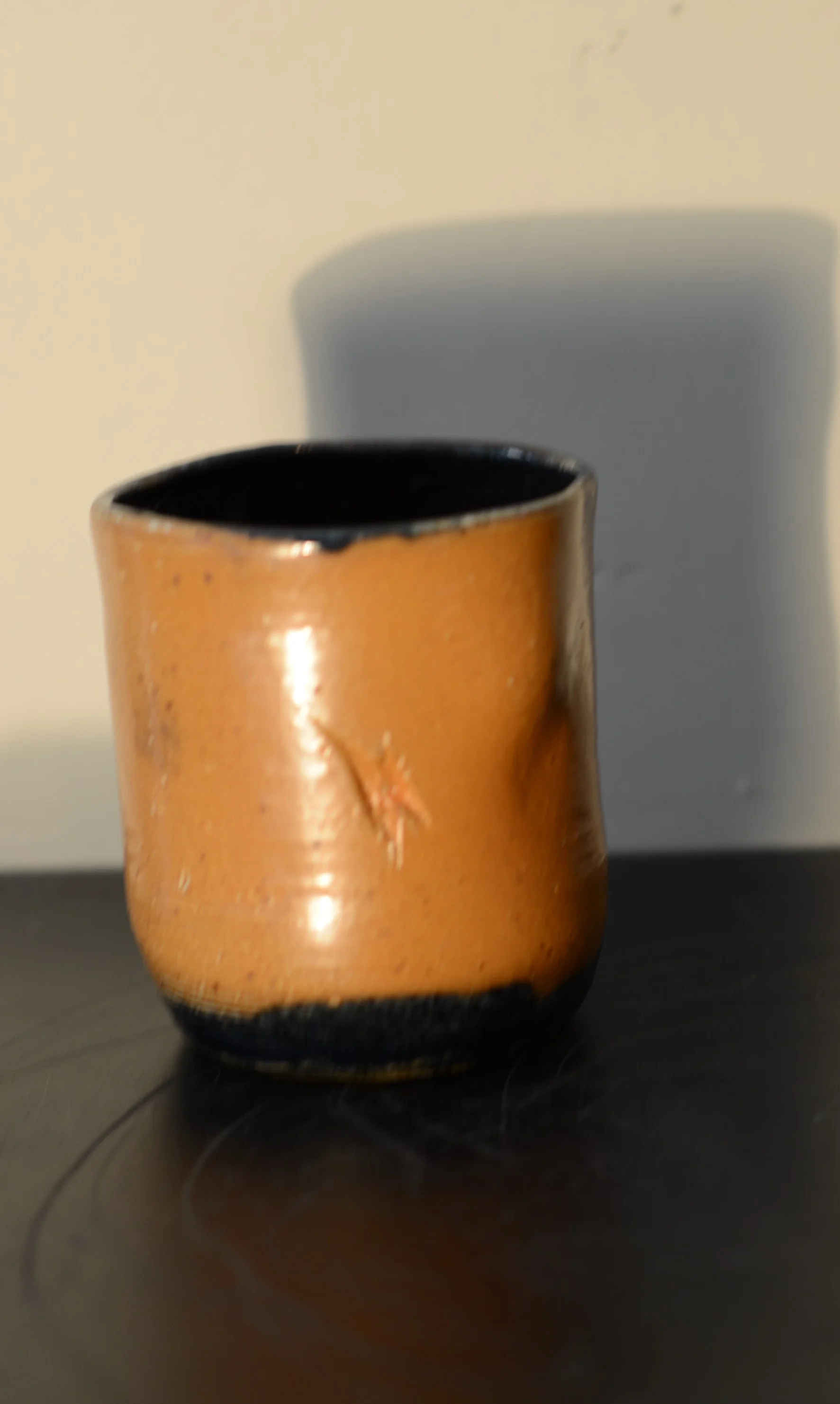 A small ceramic cup with a glazed orange exterior and black rim, placed on a dark surface with a white background and shadow.