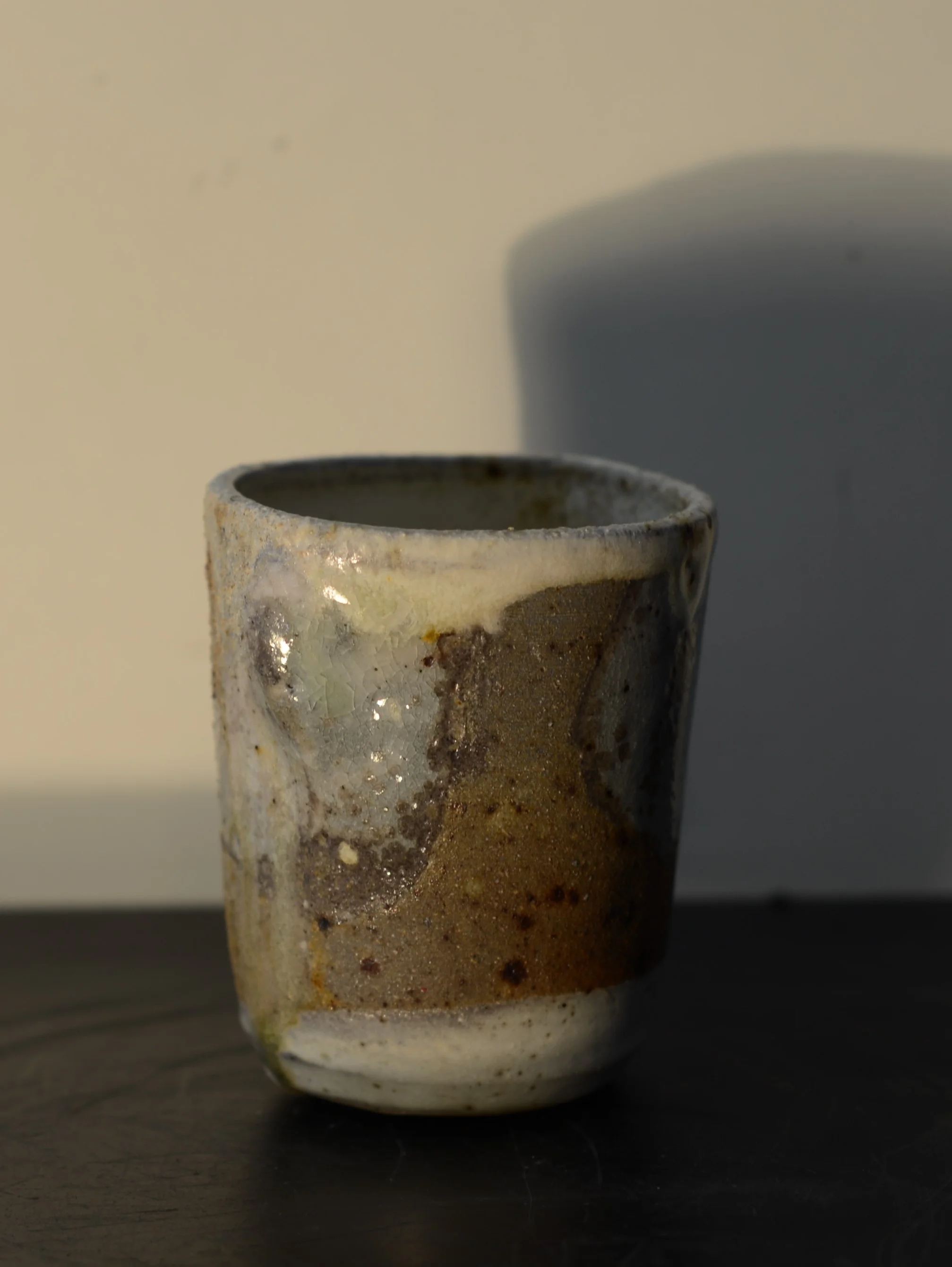 A small, cylindrical ceramic cup with a textured, uneven glaze in earthy tones of white, brown, and green, sitting on a dark surface with a shadow cast on a plain light background.