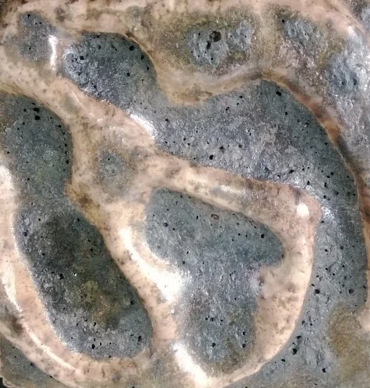 Close-up of a metallic or mineral surface with textured, irregular patterns and small black dots.