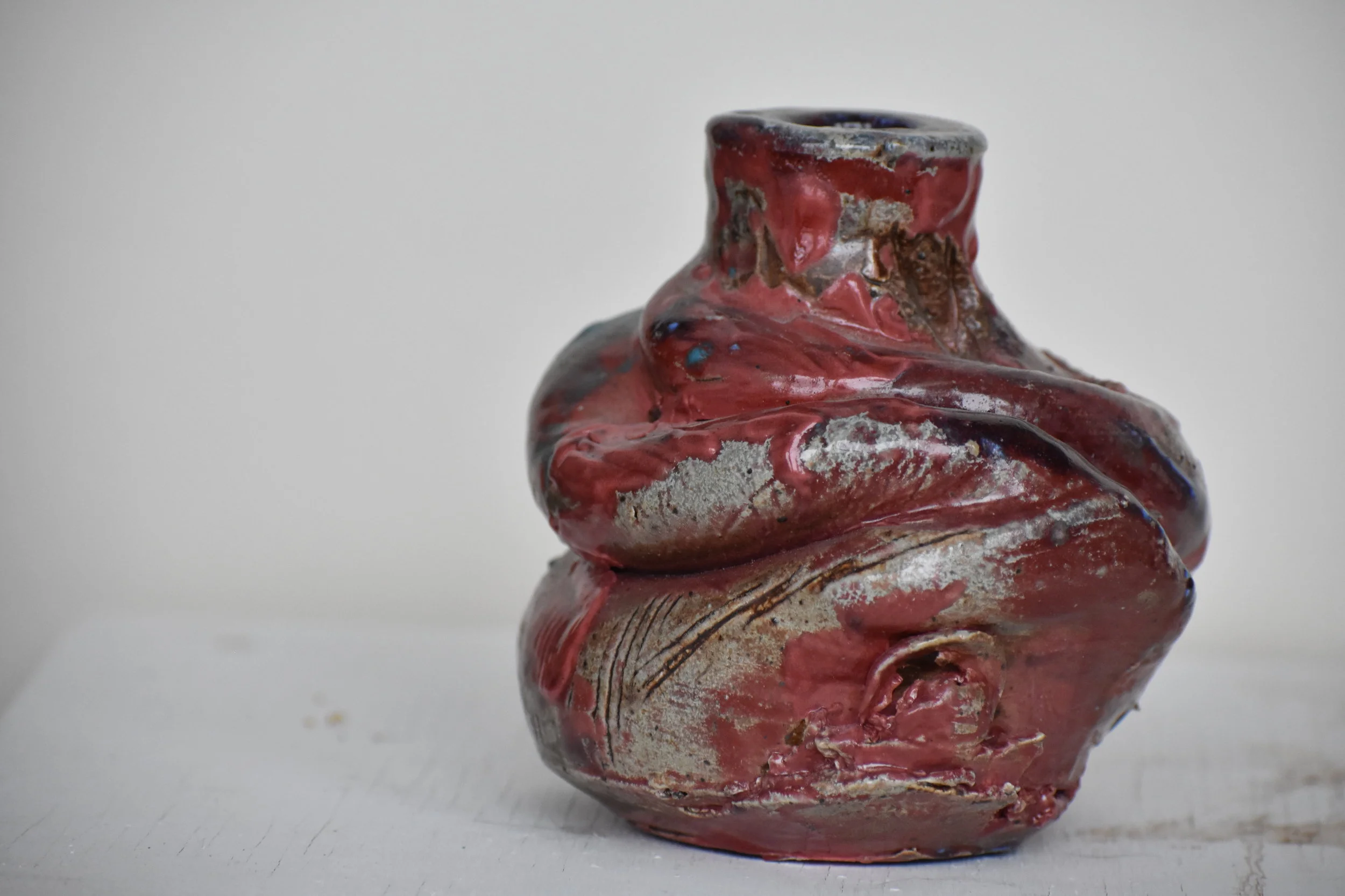 Close-up of a small, ancient, weathered ceramic vase with a reddish and grayish glazed surface, placed on a white surface against a plain white background.