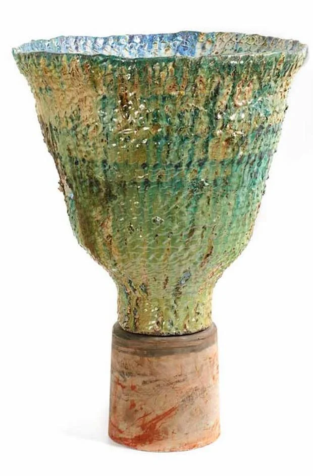 A ceramic vase with a textured, green and blue glazed surface, mounted on a short, reddish-brown pedestal.