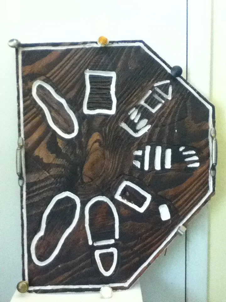 A decorative board with white outlines of various objects such as a shoe, a bottle, a fork, a spoon, a hand, and a paperclip, mounted on a wooden surface.