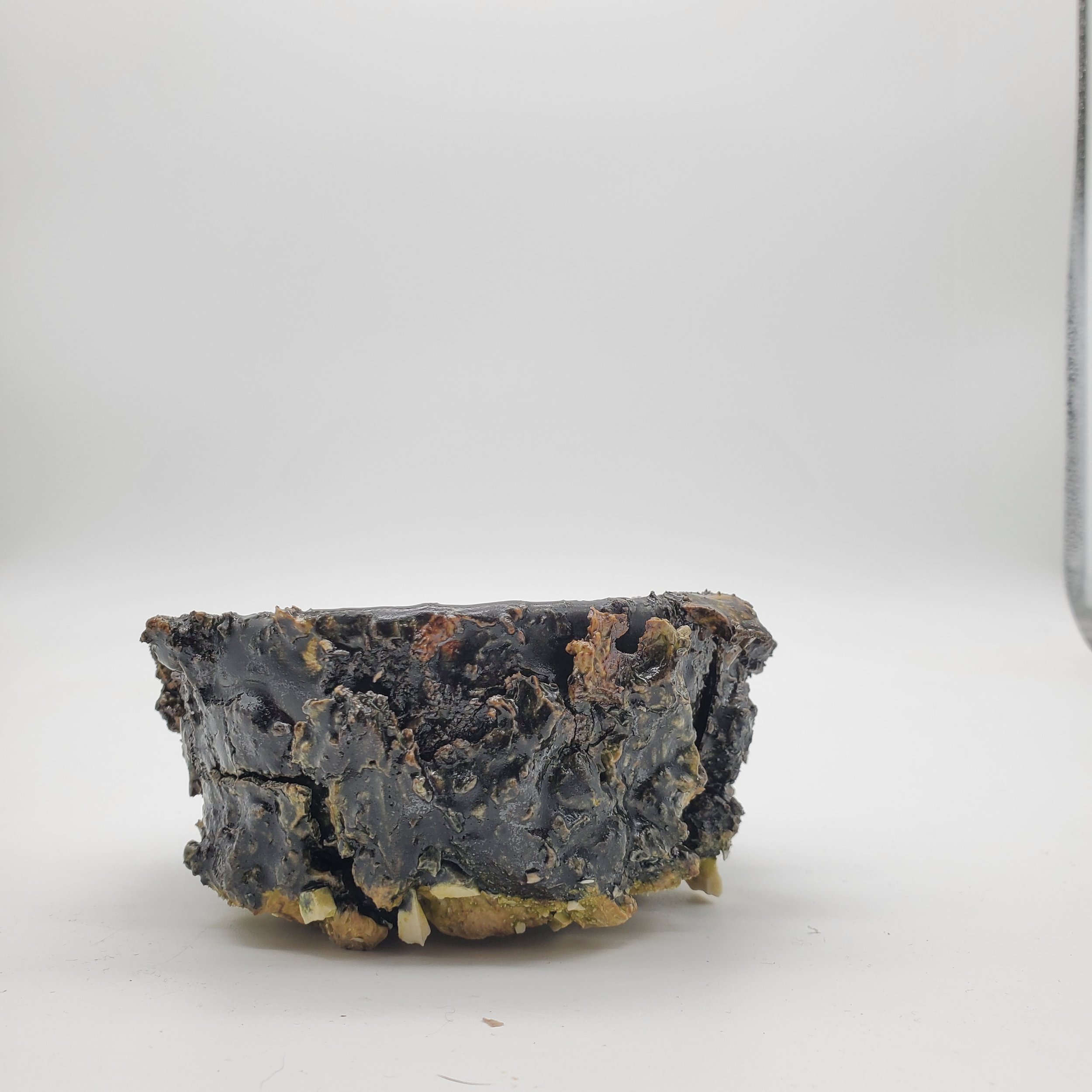 Close-up of a large, rough, black charred object with some yellowish and brownish patches, placed on a white surface against a plain white background.