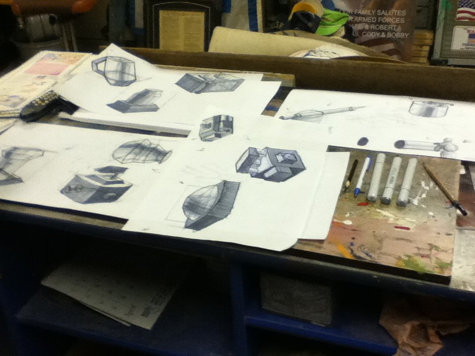 Sketches and drawings of various 3D geometric shapes and objects spread across a cluttered work table, with markers, pens, and paper.