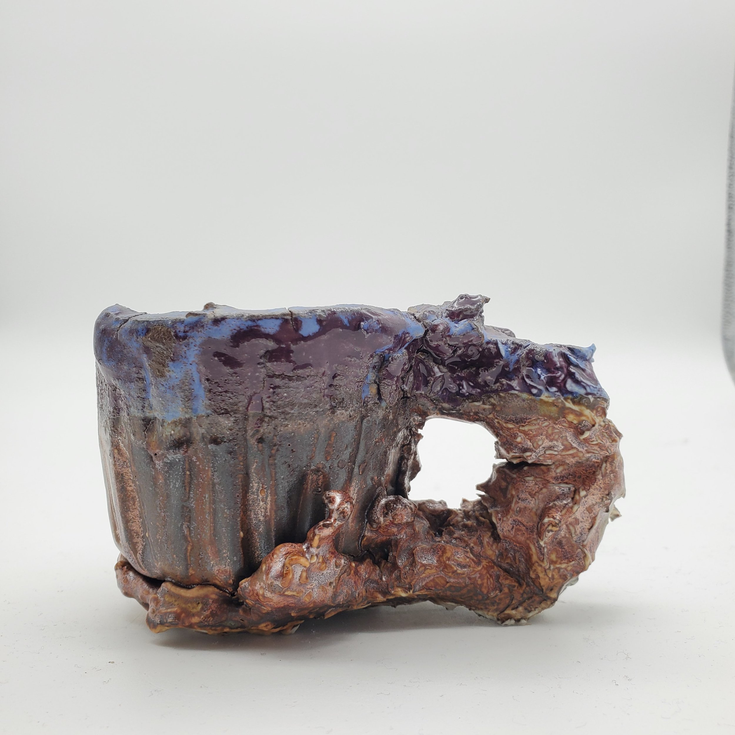 A close-up photograph of a ceramic mug with a handle, featuring an abstract, textured glaze in shades of purple, blue, and brown, set against a plain white background.