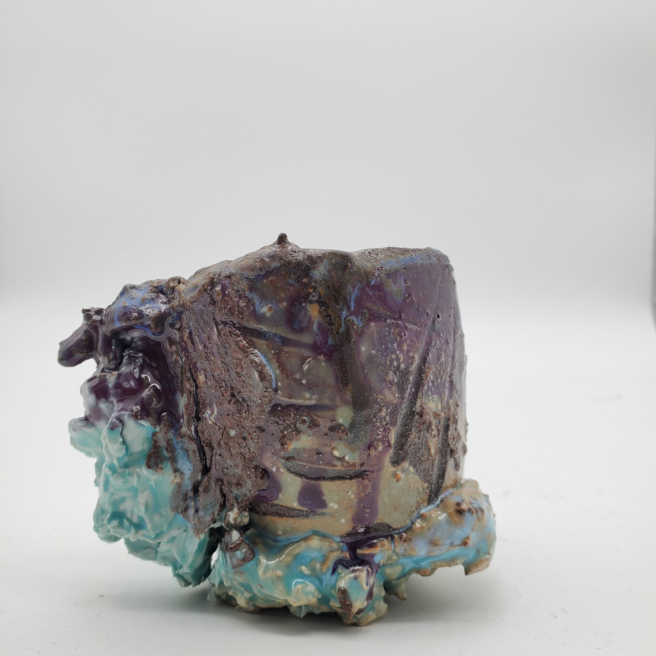 Close-up of a ceramic art piece or sculpture with a rough texture, colorful glaze, and abstract form, placed on a plain white background.