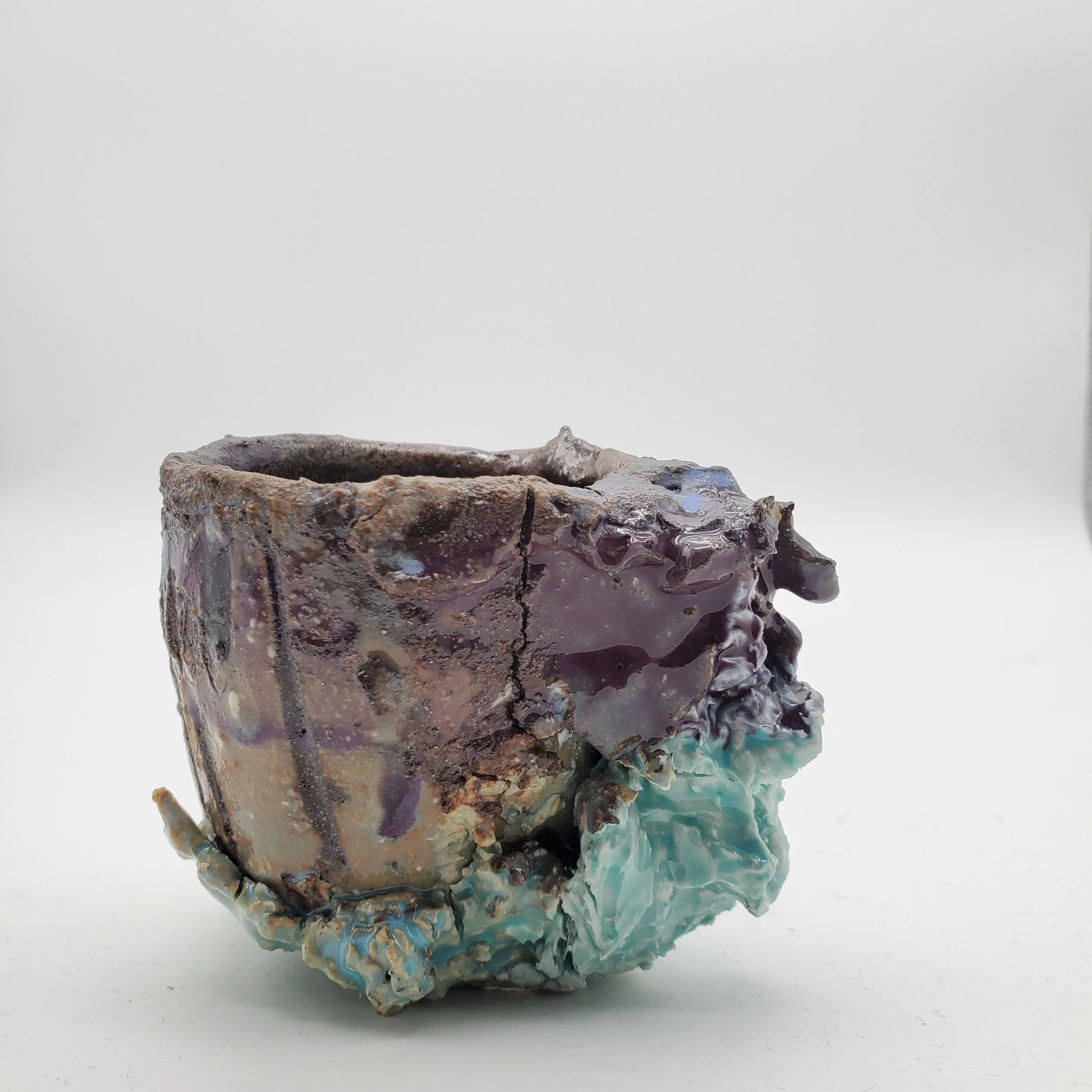 A ceramic art piece resembling a cup made with textured, multicolored glaze and an irregular, organic shape on a plain white background.