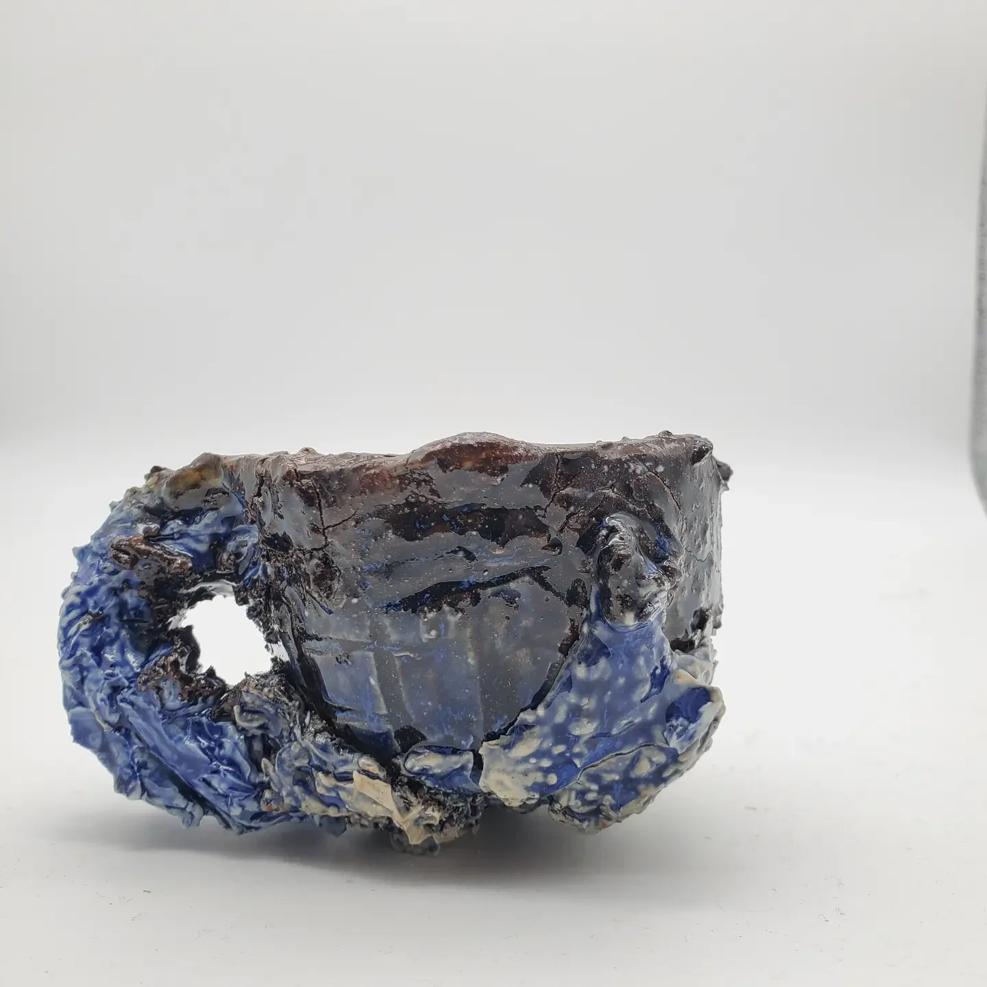 A close-up of a melted, charred ceramic mug with a blue and white glaze, placed on a white surface with a plain white background.