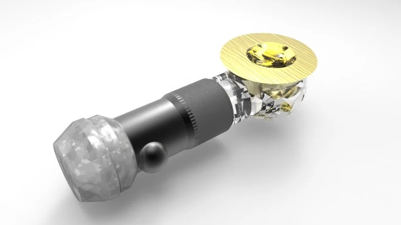 Close-up of a small mechanical device with a gold-colored circular component on one end, a black cylindrical section in the middle, and a faceted transparent section, resting on a plain white surface.