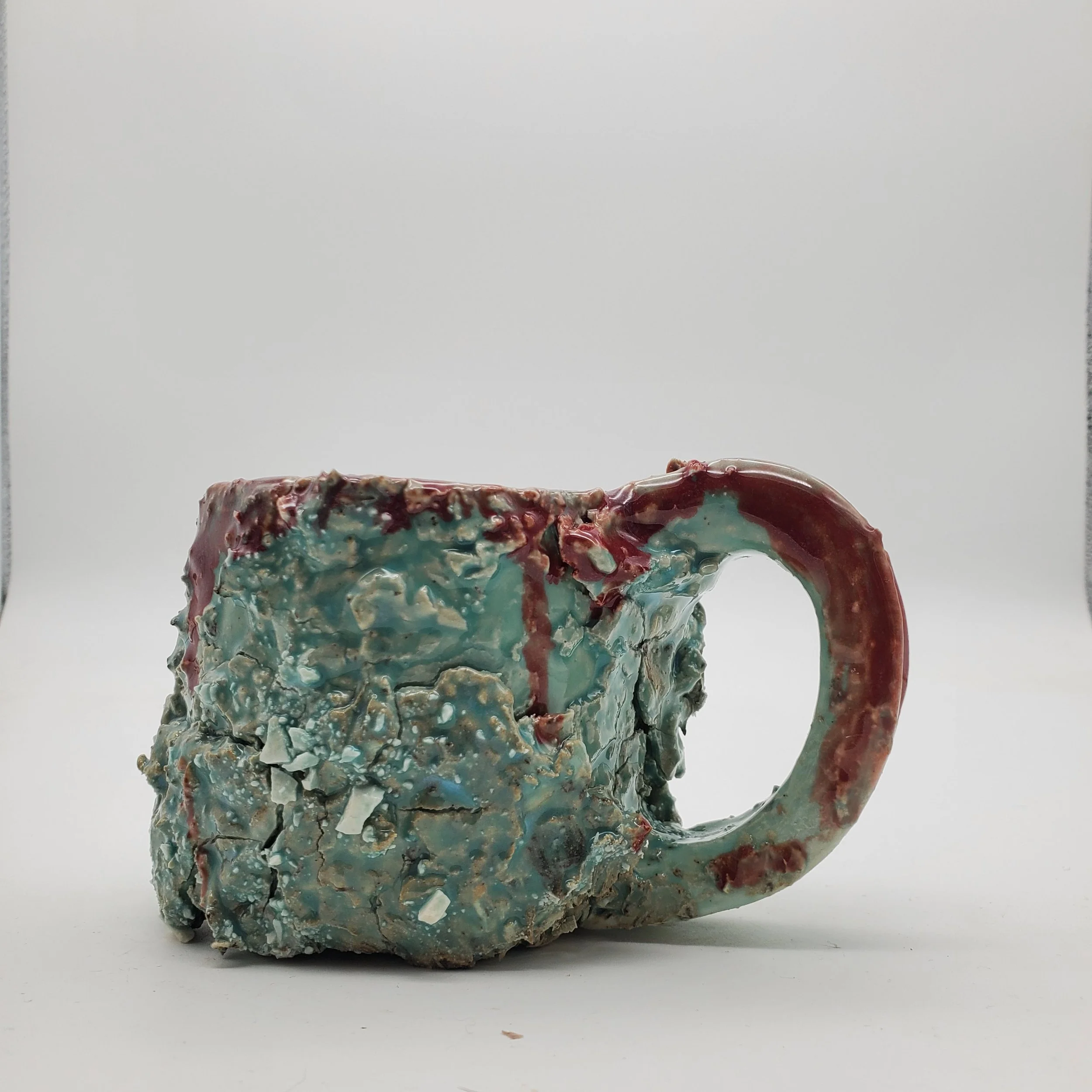 A weathered, cracked ceramic mug with chipped glaze, featuring a blue, turquoise, and red color palette, set against a white background.