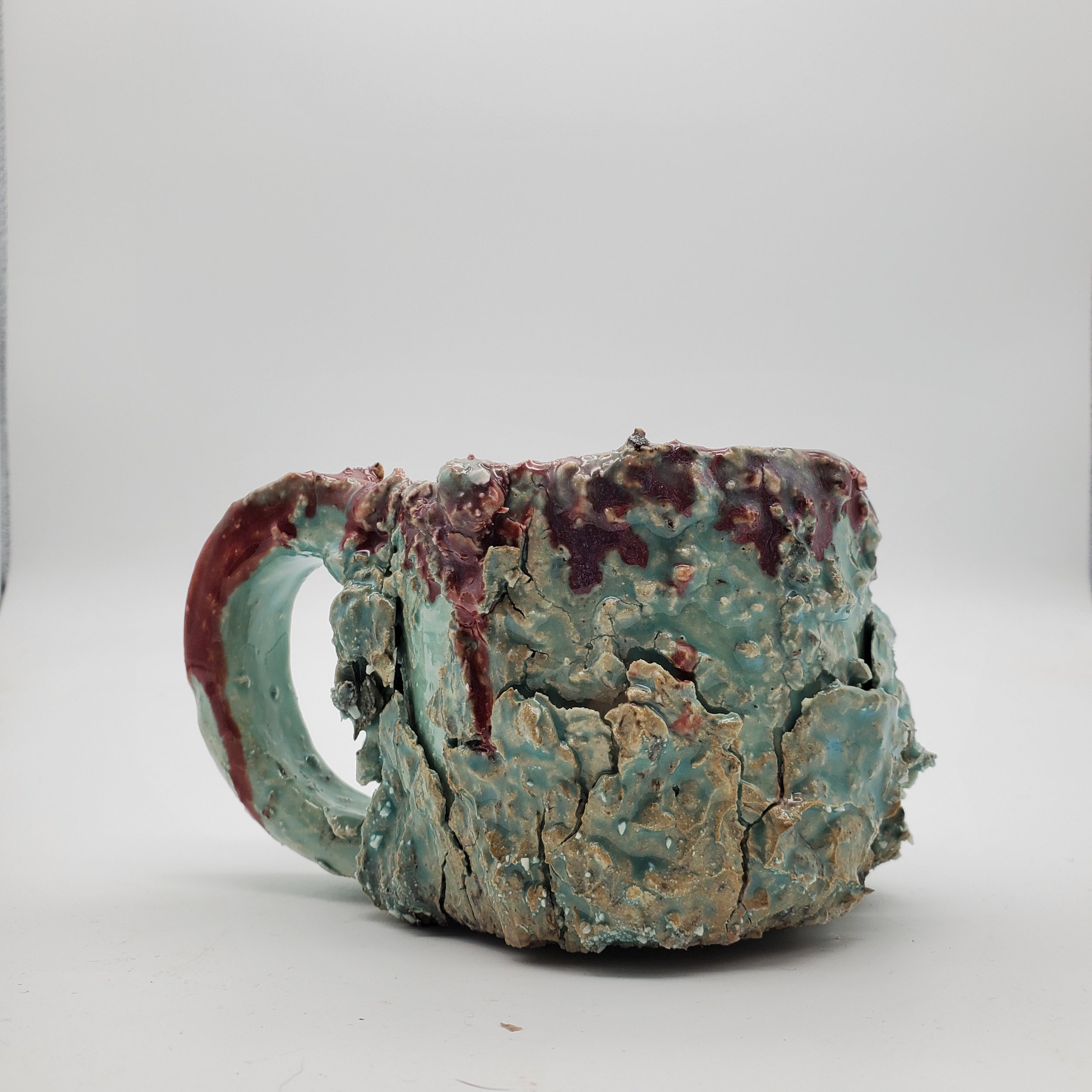 A ceramic mug with a textured, crackled surface in shades of turquoise, brown, and red, placed against a plain white background.