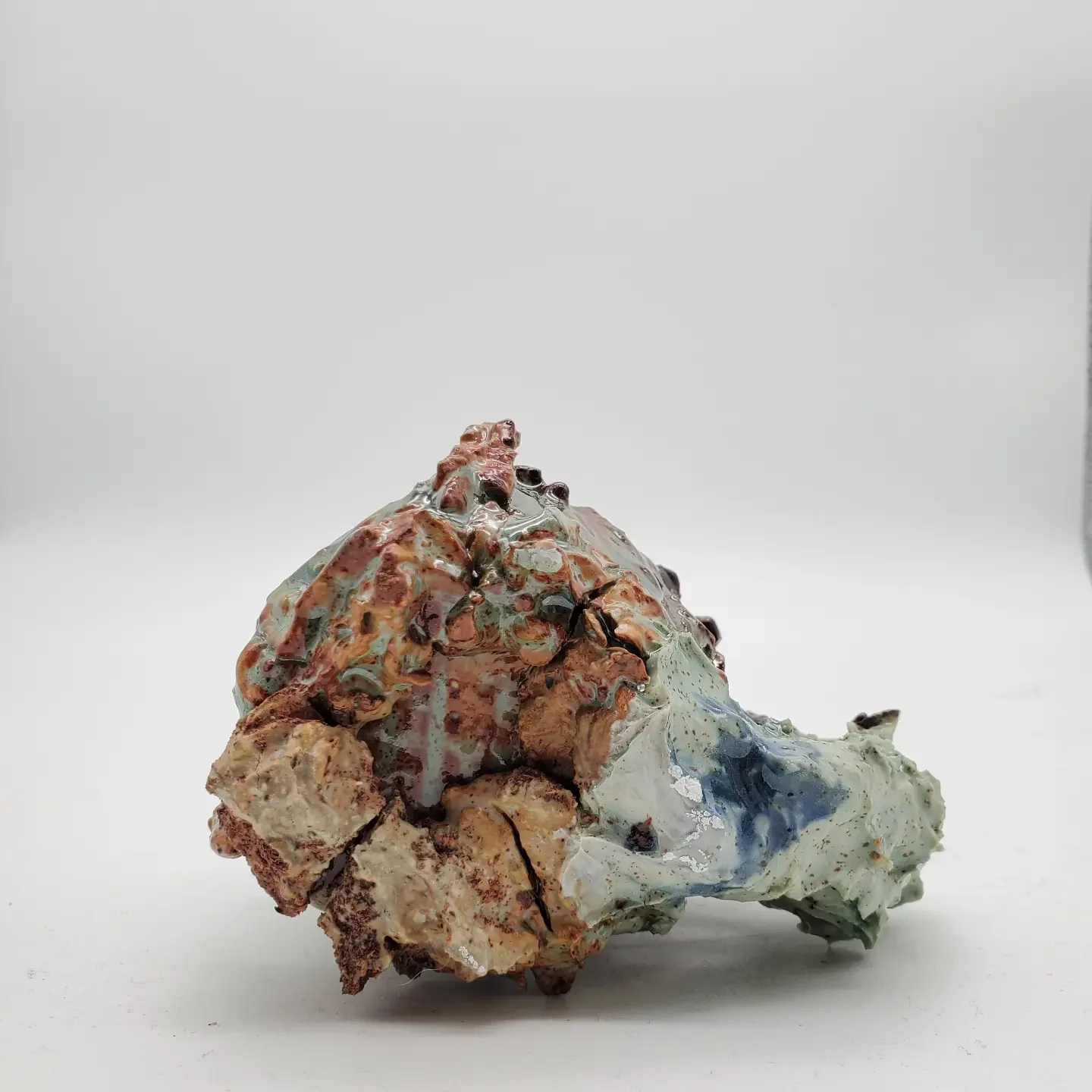 A ceramic sculpture resembling a geological formation or an abstract mountain, with textured, uneven surfaces and multiple layered colors including browns, greens, blues, and whites, set against a plain white background.