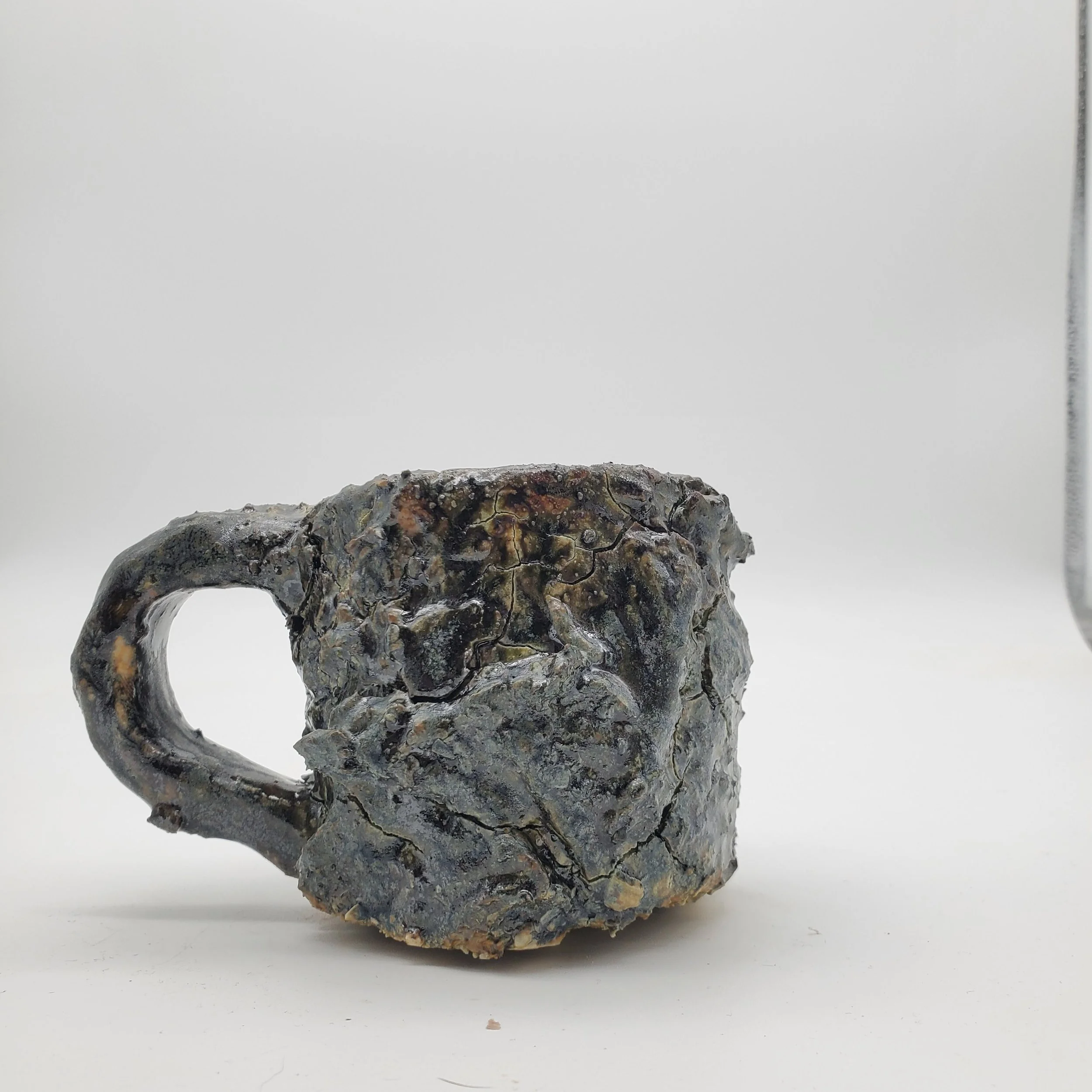 A mug made from a rough, cracked, and textured metal-like material on a white surface with a plain background.