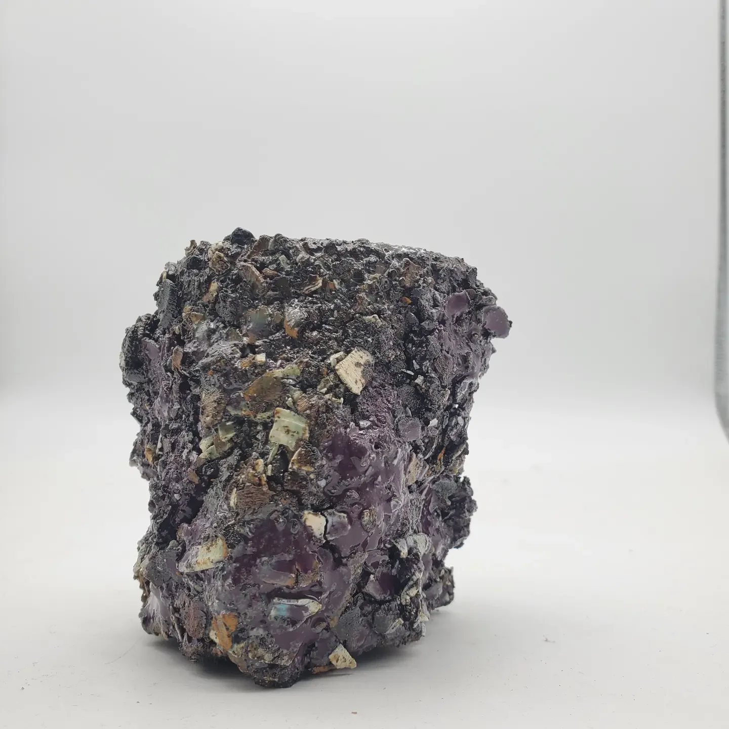 A rough, irregularly shaped, dark purple mineral specimen with small, shiny, metallic-looking mineral fragments embedded in it, displayed against a plain white background.