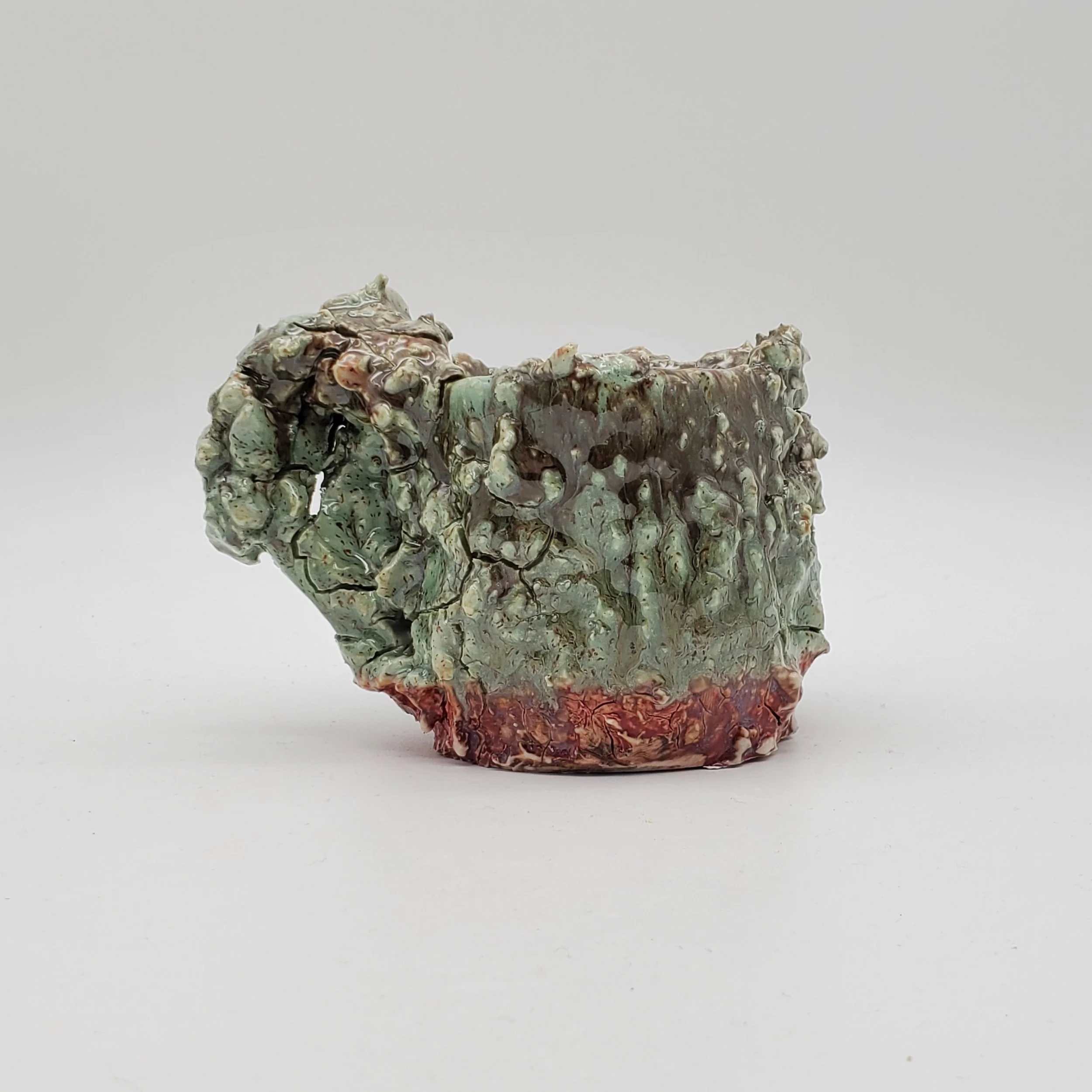 A small ceramic sculpture with a textured, irregular surface featuring green, brown, and reddish-orange glaze, on a plain white background.