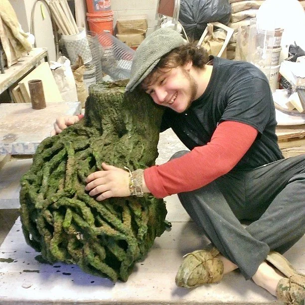 A young man with dark hair, wearing a gray hat, black shirt with red sleeves, and gray pants, is sitting on the floor and hugging a large moss-covered piece of driftwood or sculpture, smiling happily. The background shows a workshop with various tool