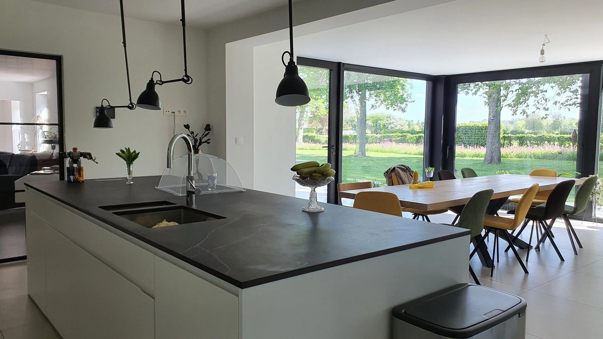 Modern kitchen with white cabinetry, black countertop, and hanging black pendant lights, adjacent to a dining area with a wooden table and multicolored chairs, large windows with a view of a green outdoor landscape.