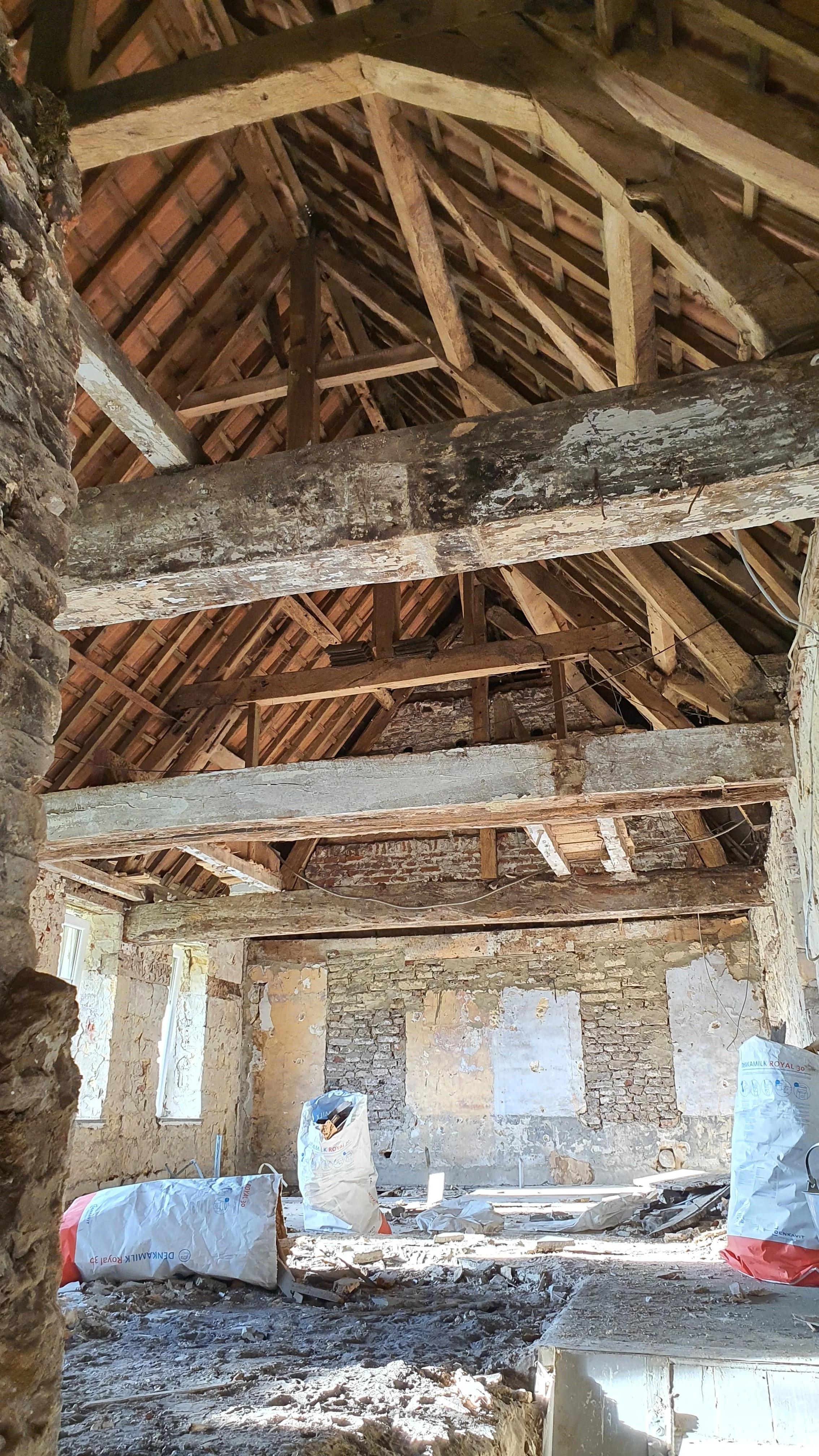 Interior of an attic or upper story of a building under renovation, showing exposed wooden beams, brick walls, and construction materials on the floor.
