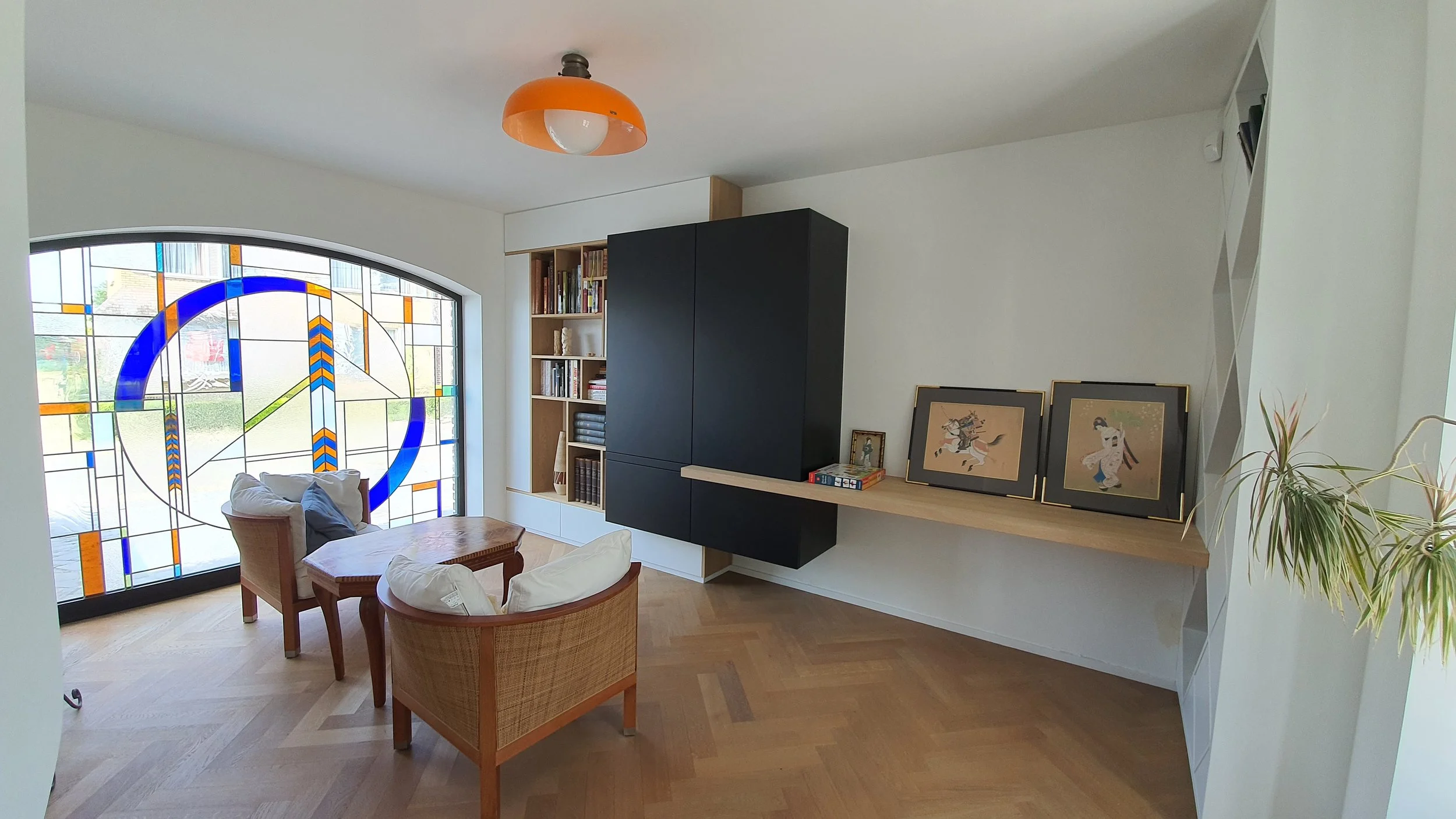 Living room with wooden furniture, two armchairs with pillows, a black and wood cabinet, a bookshelf with books, a stained glass window with a blue circle and orange details, and framed artwork on a shelf.