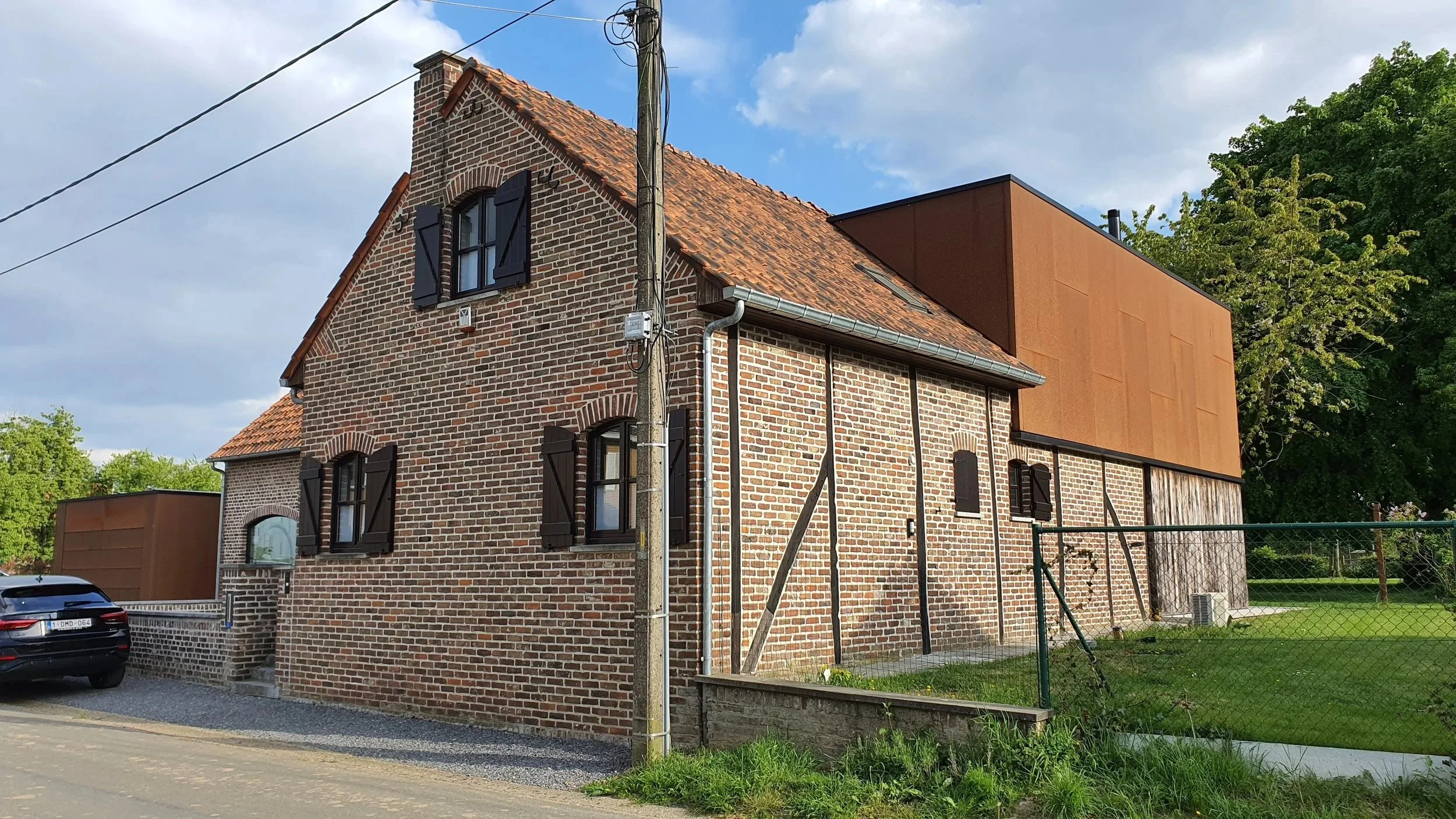 A brick house with black window shutters and a tiled roof, next to a modern wooden and cortensteel structure, with a fence and green lawn, under a cloudy sky.