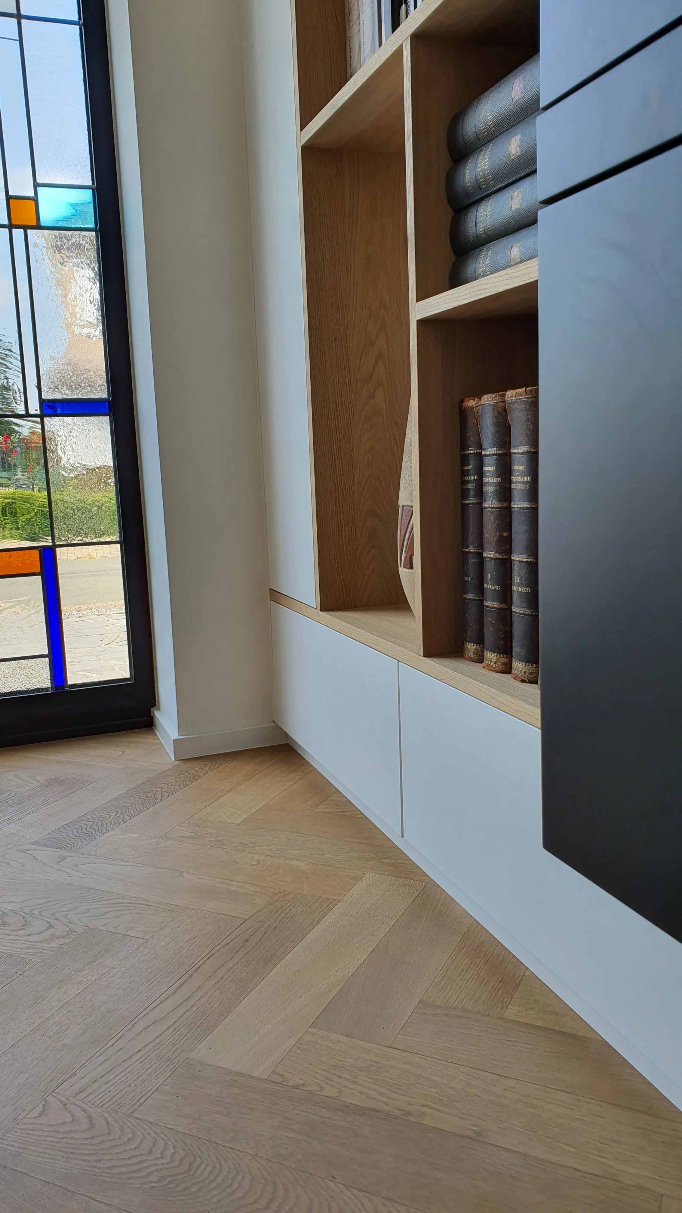 Bookcase with dark leather-bound books and modern wall with stained glass window beside light wooden flooring.