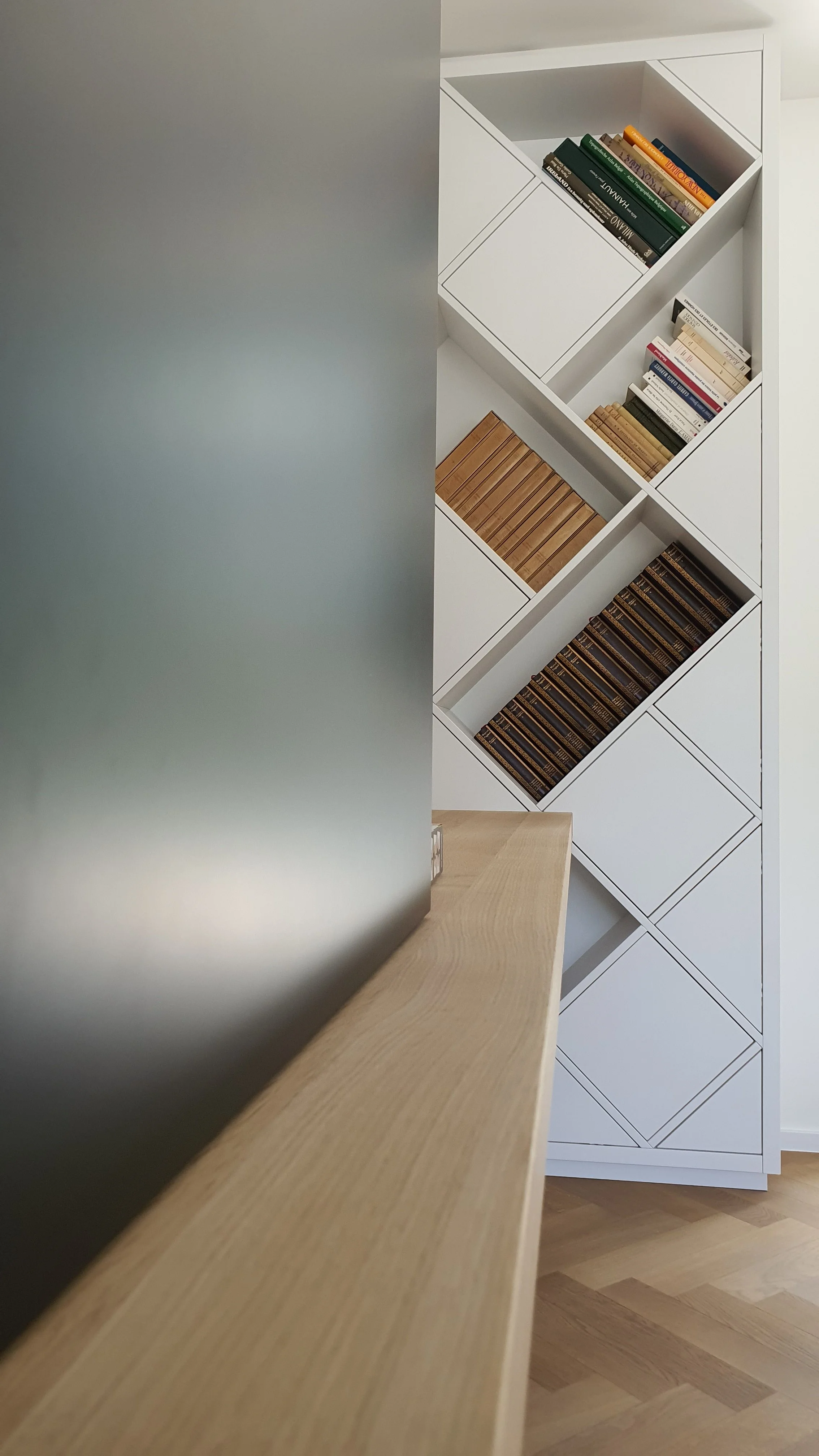 A modern white bookshelf with diagonal compartments filled with books, seen from behind a frosted glass panel and a wooden surface.