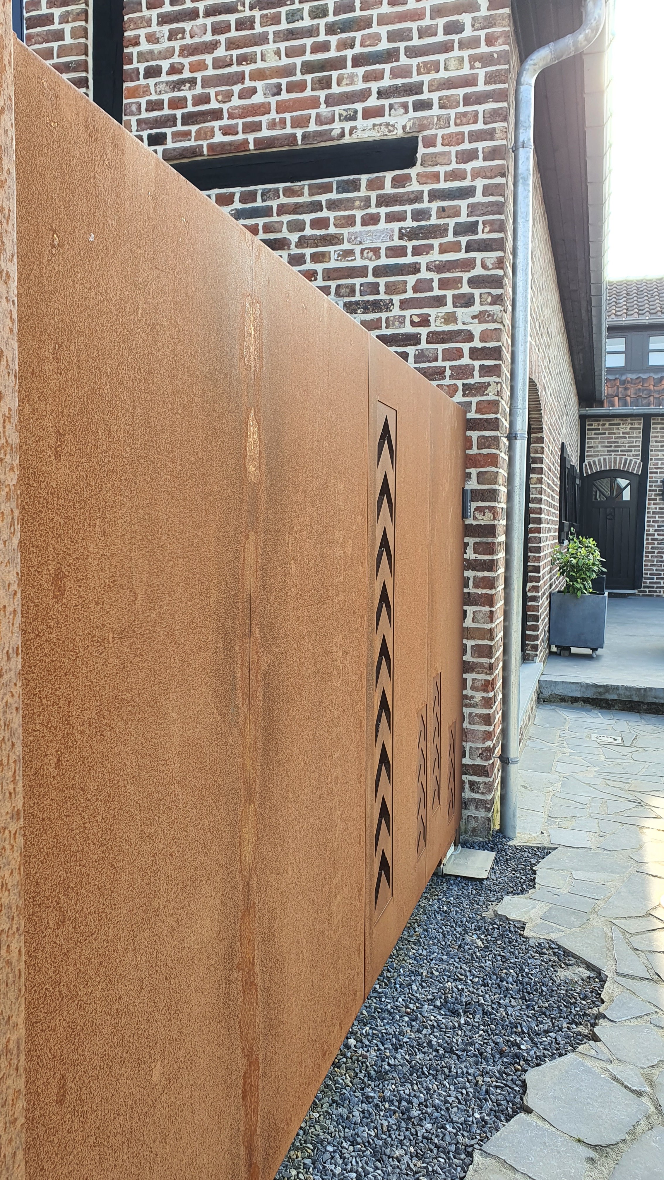 The image shows a rust-colored metal utility box with slanted ventilation cutouts, situated outdoors next to a brick wall and a drain pipe, with a stone pathway and some potted plants in the background.