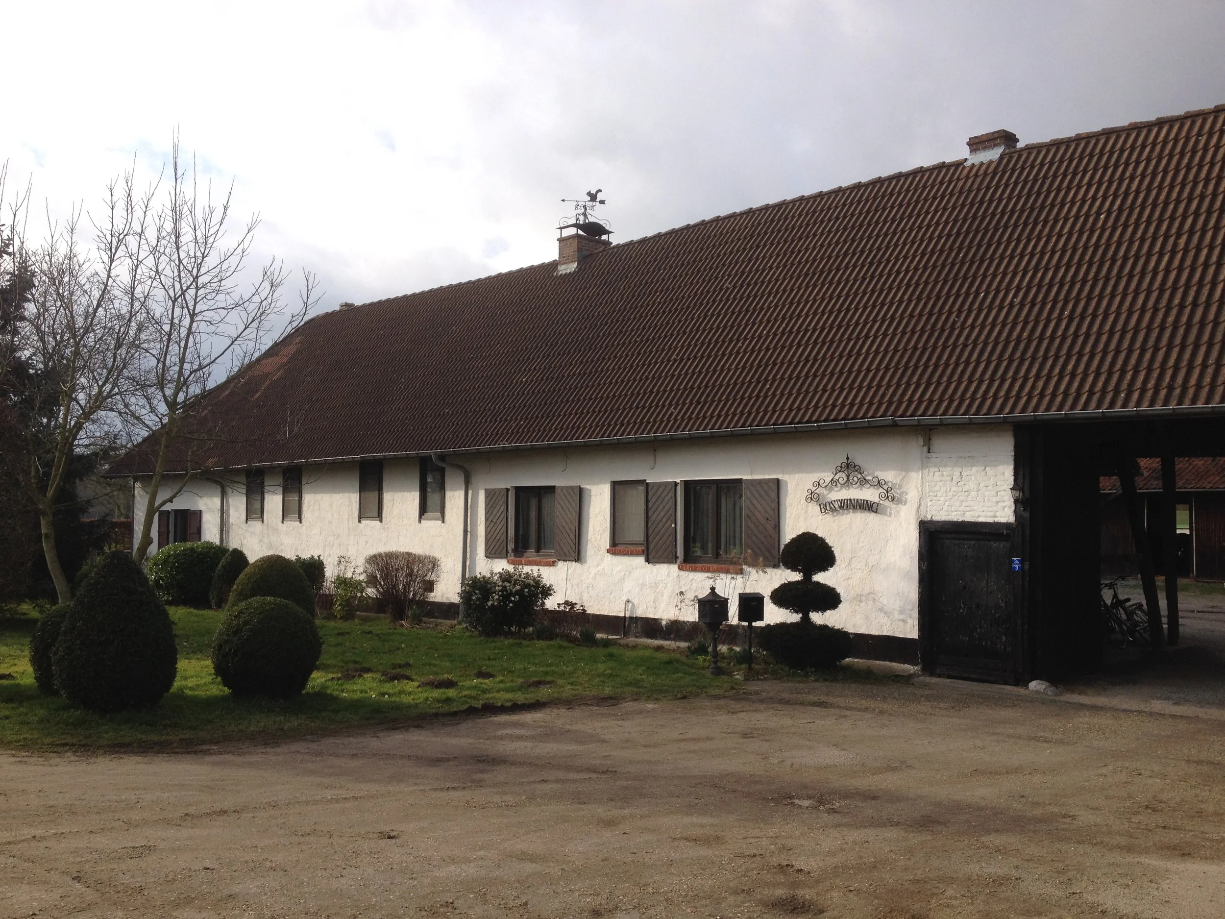 A white barn with a brown tiled roof, black window shutters, and a sign that reads "Boswinning." The barn is surrounded by a lawn with neatly trimmed bushes, some trees, and a dirt driveway.