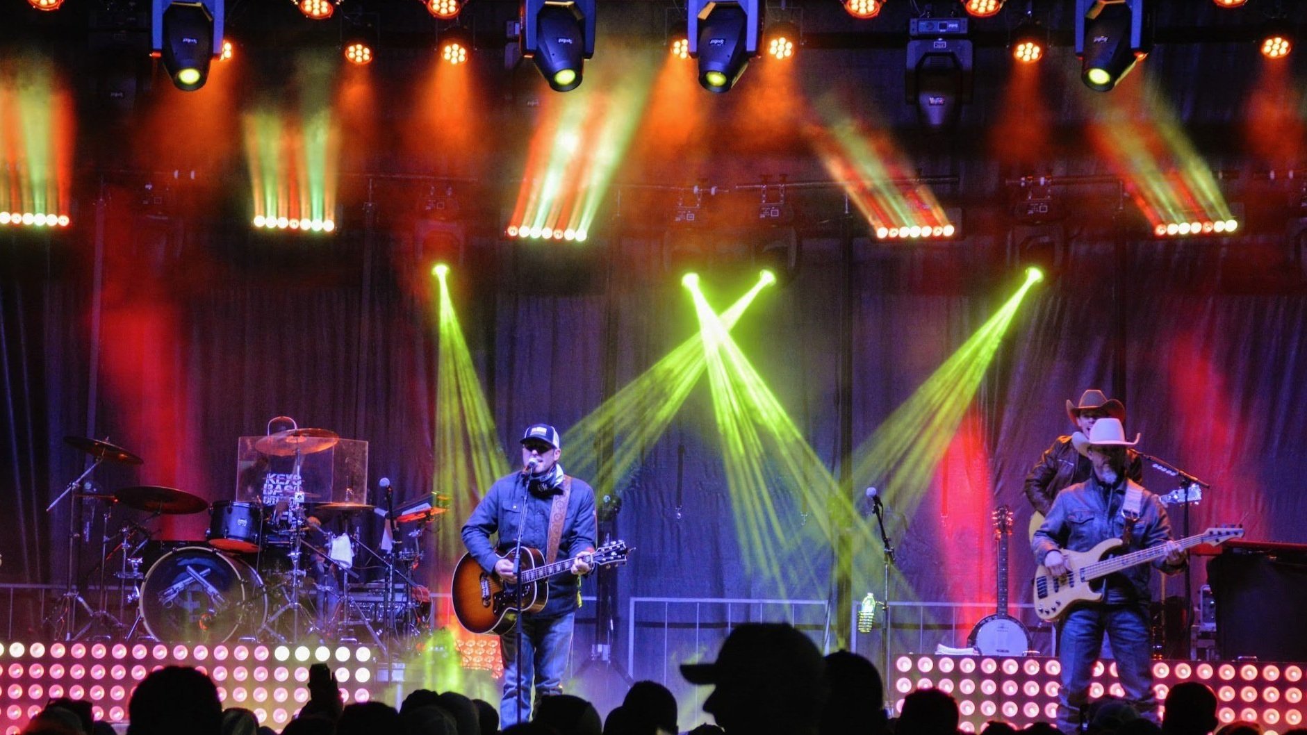 Musicians performing on stage with colorful lighting and silhouettes of audience in front.