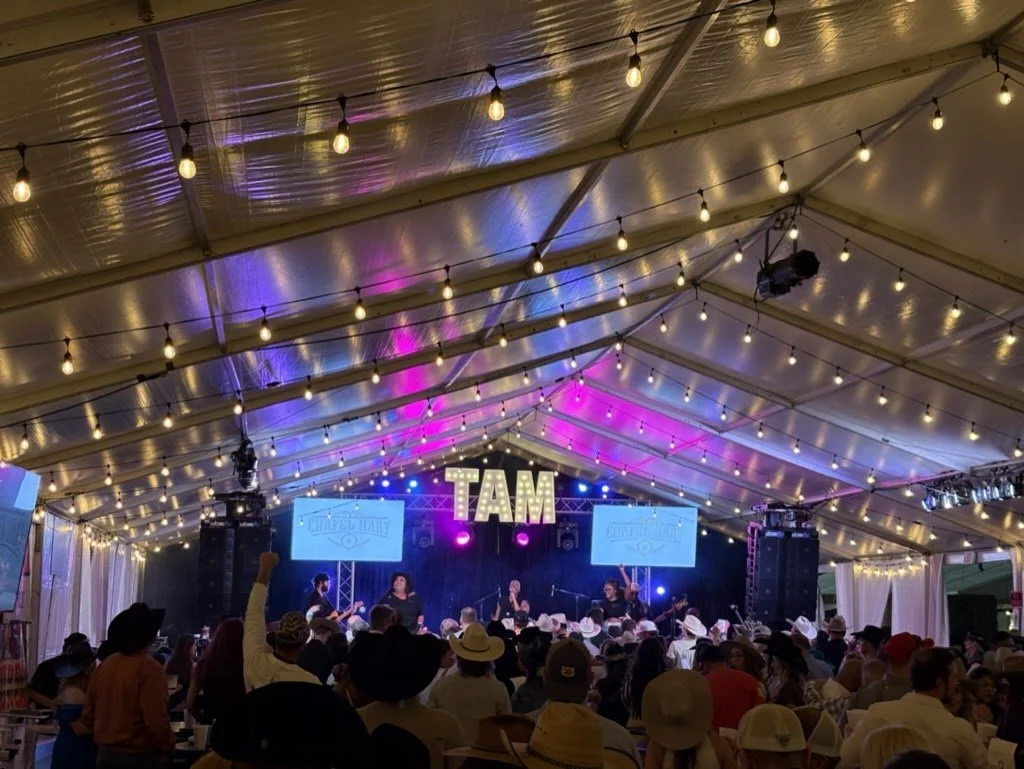 Music concert inside a large tent with string lights, a band performing on stage, and large illuminated letters spelling "TAM" hanging above.