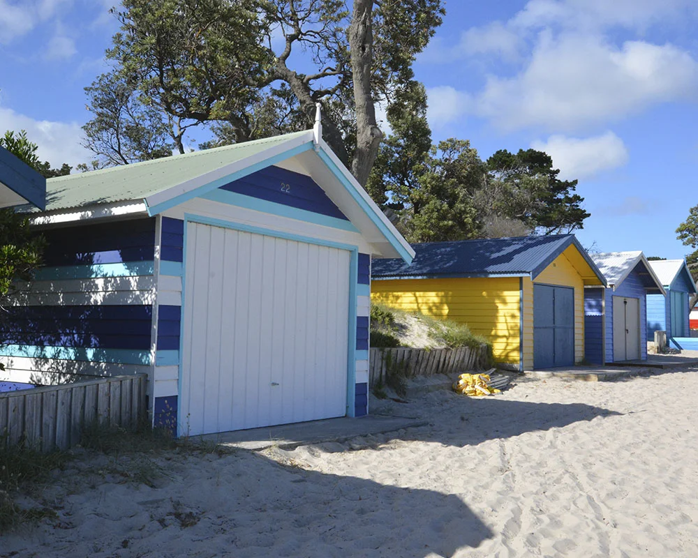 Colorful beach huts with closed doors on sandy terrain under a partly cloudy sky.
