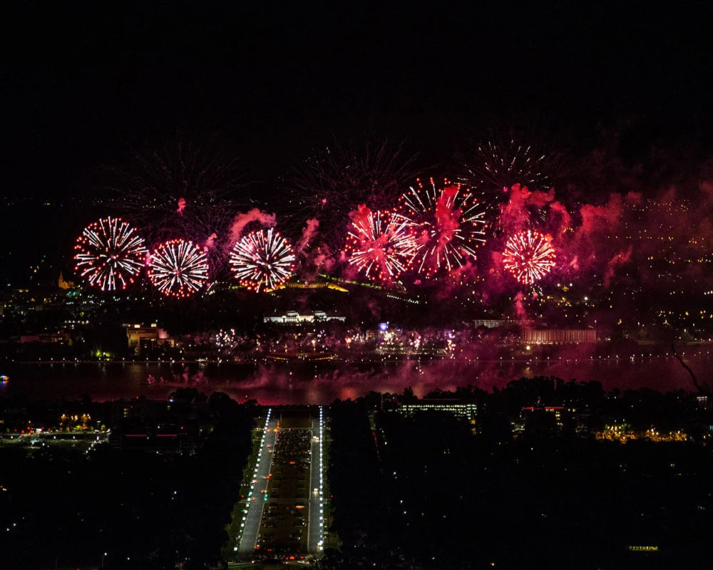 Fireworks display over a city at night, with colorful explosions illuminating the sky, near a river and cityscape. Canberra Skyfire event.