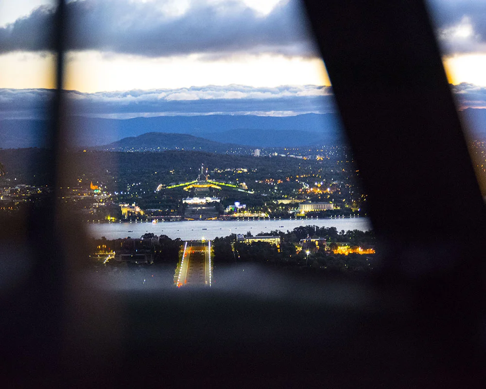 View of a cityscape at dusk, with a river, bridge with lights, and mountains in the background, seen through window blinds.