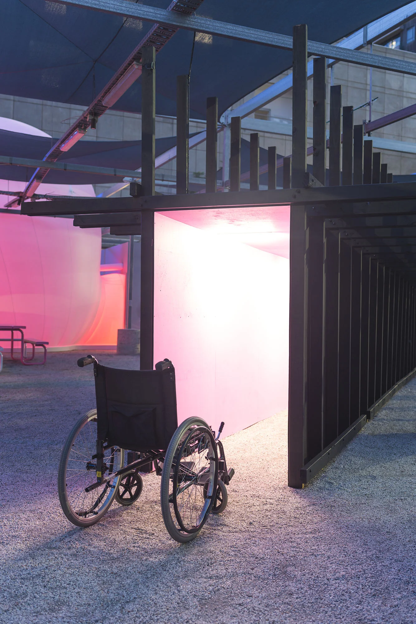 Install view. A wheelchair sits outside at night in front of a corridor, painted pink and lit from within by horizontal neon lights. The skeleton of the corridor is exposed and painted black.