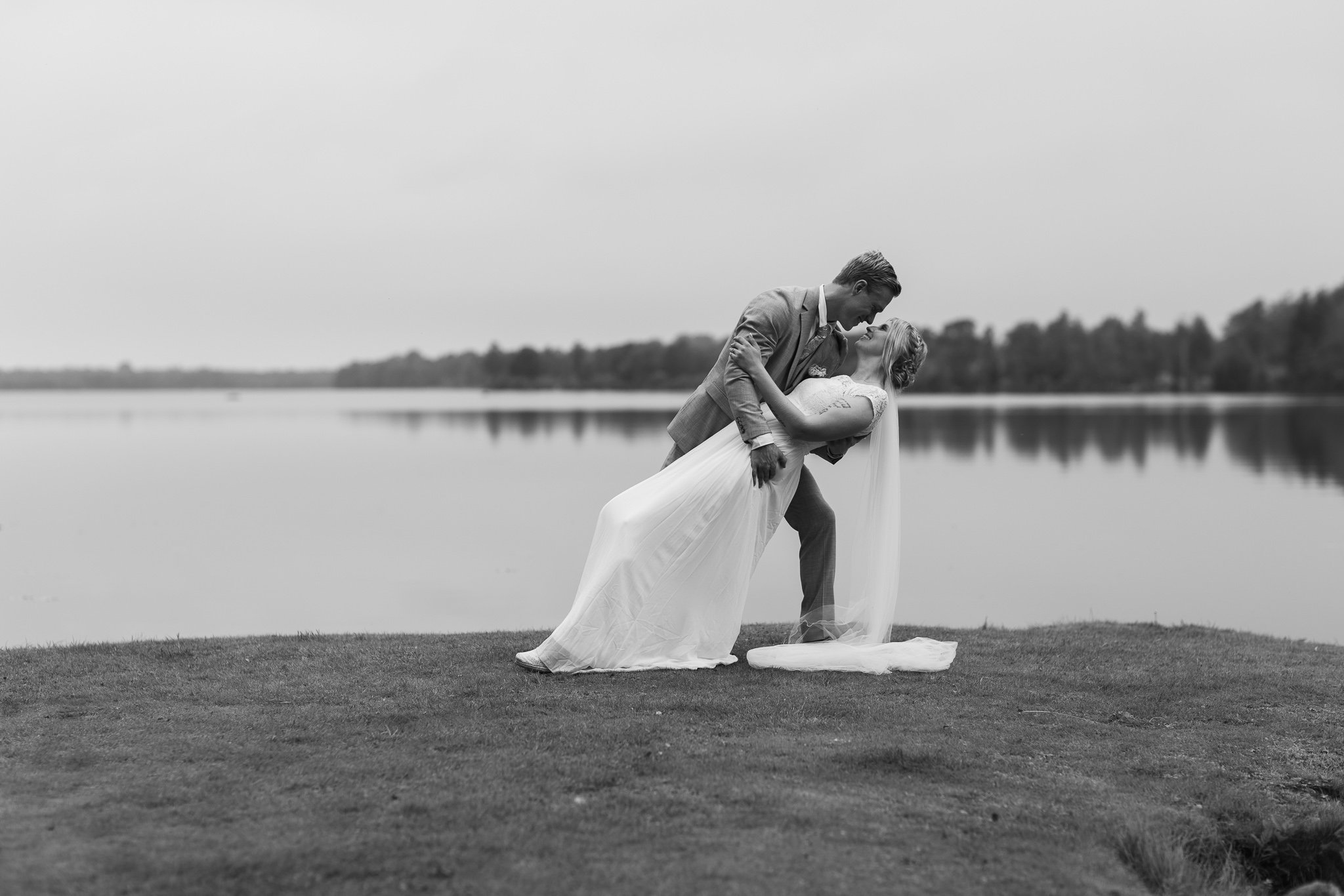 En svartvit bild av ett brudpar utomhus vid en sjö, där mannen räddar kvinnan som lutar sig bakåt i en dansartad rörelse.