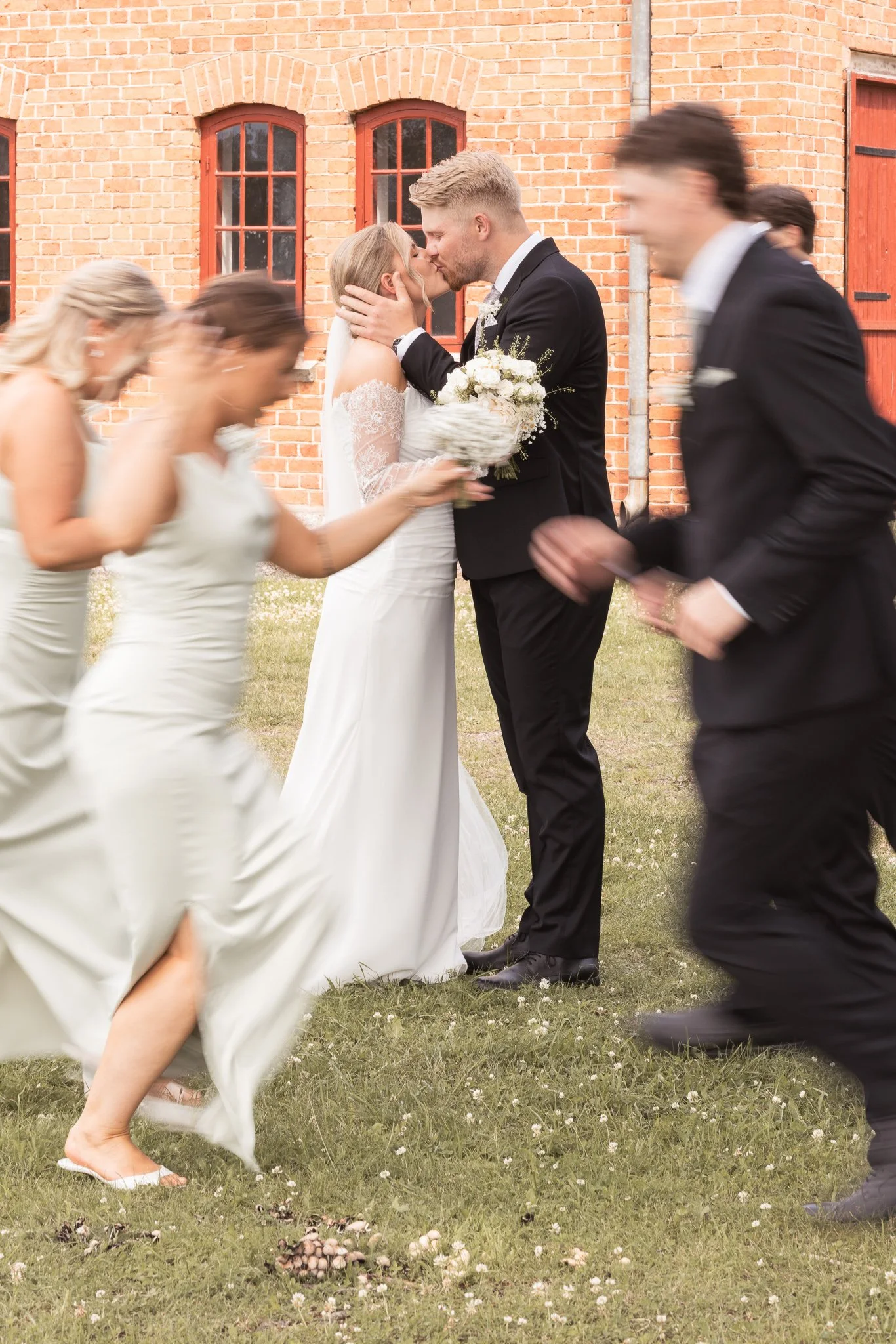 Ett brudpar kysser varandra under en bröllopsceremoni utomhus, omgiven av bröllopsgäster som dansar och leker på gräset.