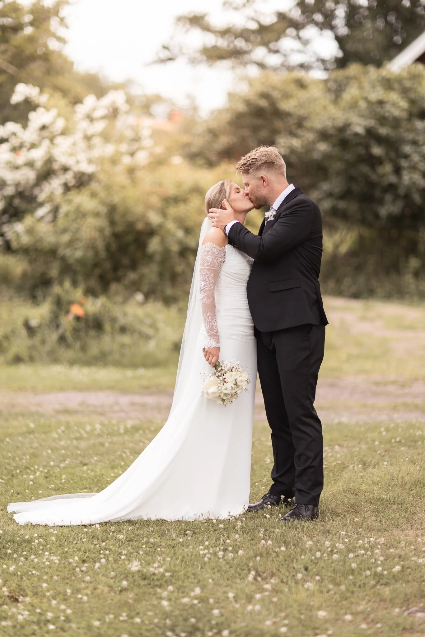 En nygiften man och kvinna kyssar varandra utomhus på en gräsmatta med blommor i bakgrunden, kvinnan håller en bukett med vita blommor.