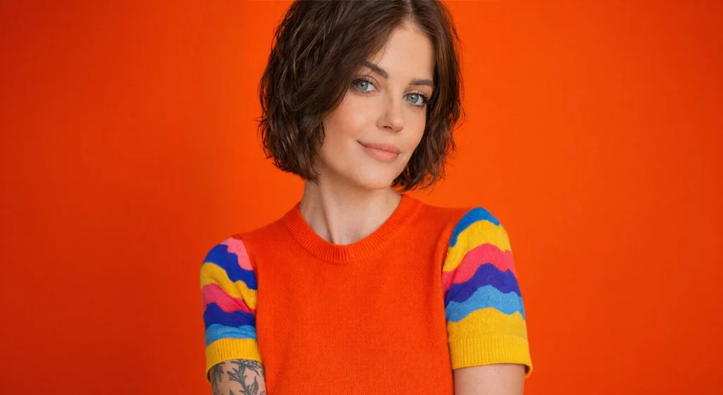 Portrait of non-binary designer Adrienne Jane wearing an orange sweater with colourful sleeves against a bright orange background.