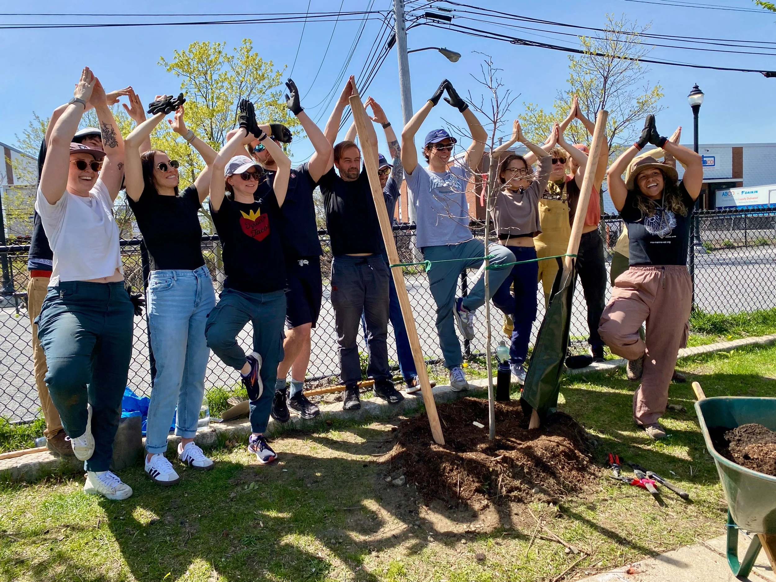 Volunteers strike a tree pose while standing behind a newly planted tree.