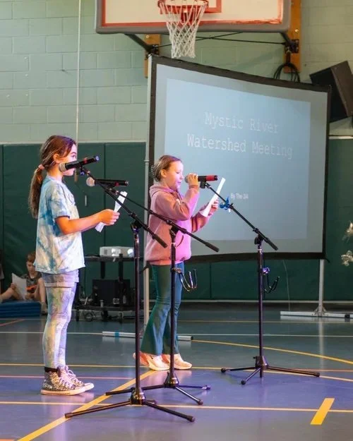 Two students speak into microphones with a slideshow in the background.