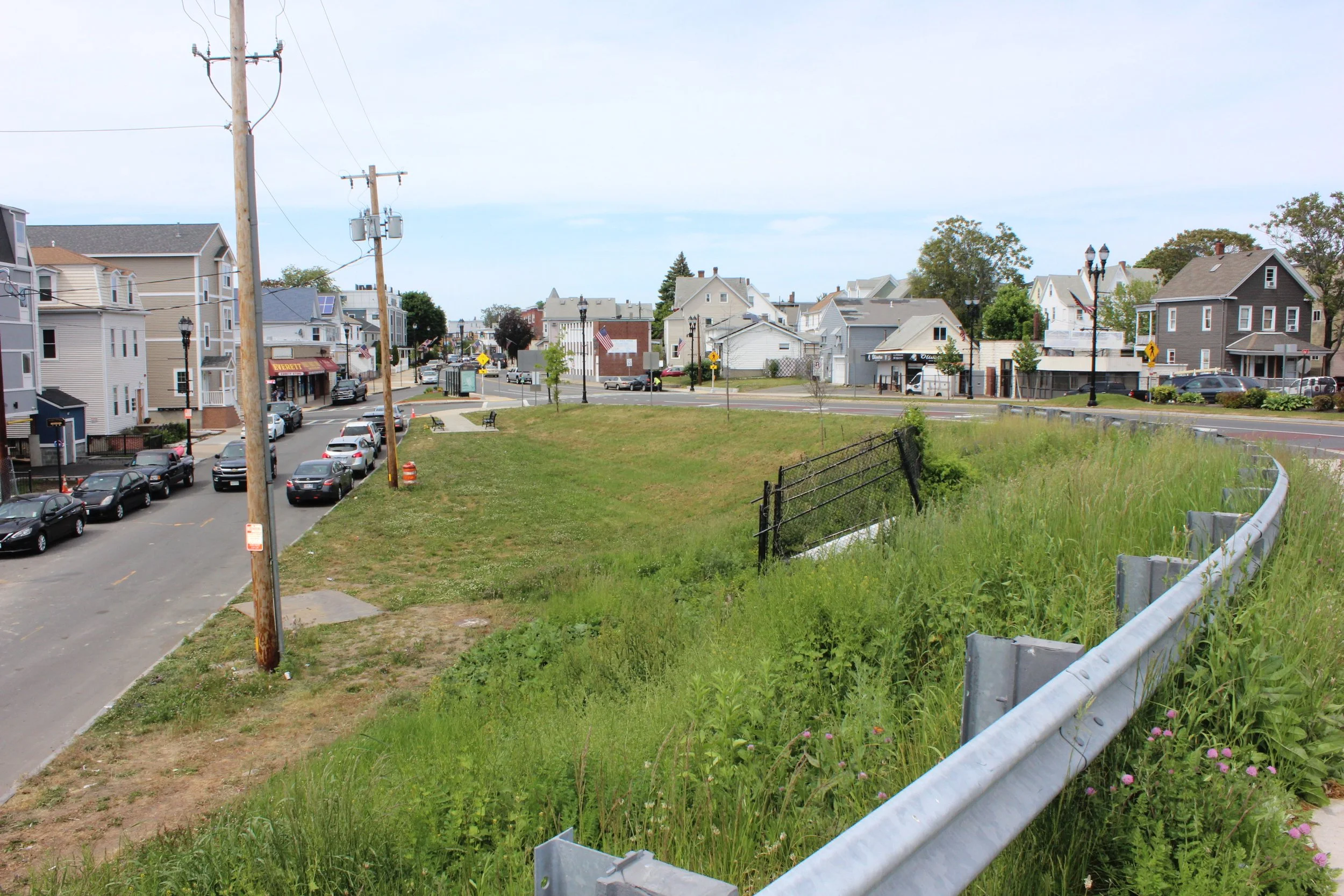 Vacant grassy lot beside a busy roadway with surrounding homes and limited tree cover.