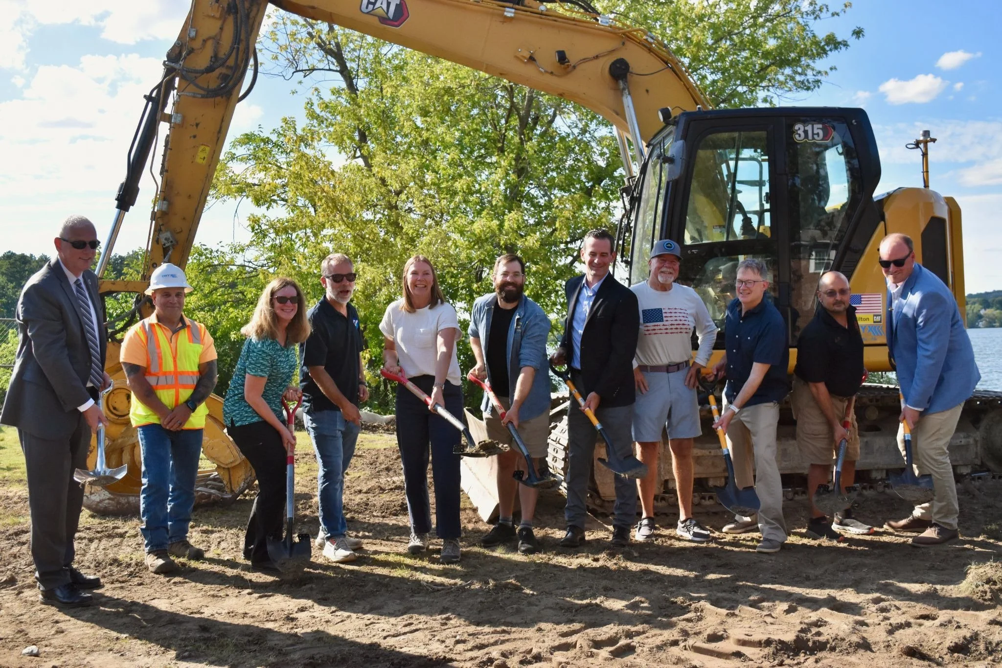 A group of 11 people hold shovels in front of construction equipment