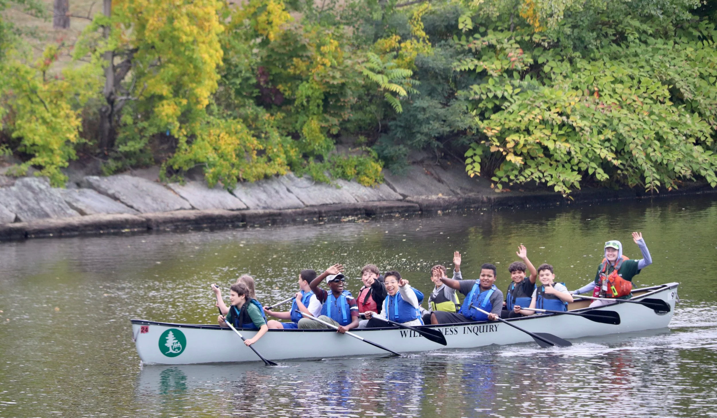 10 students and one lead paddler paddle on the Mystic River while waving at the camera