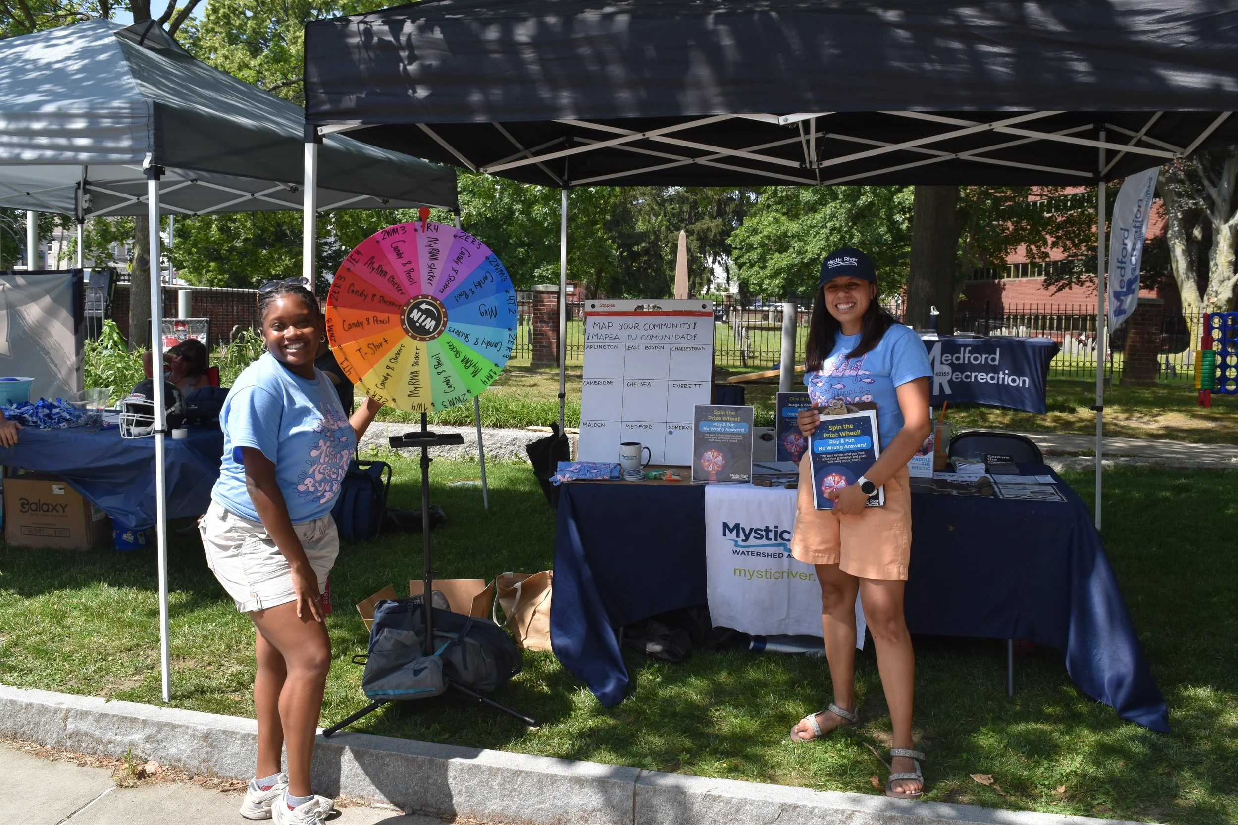 Reece and Alessandra stand by a prize wheel and an outreach table.