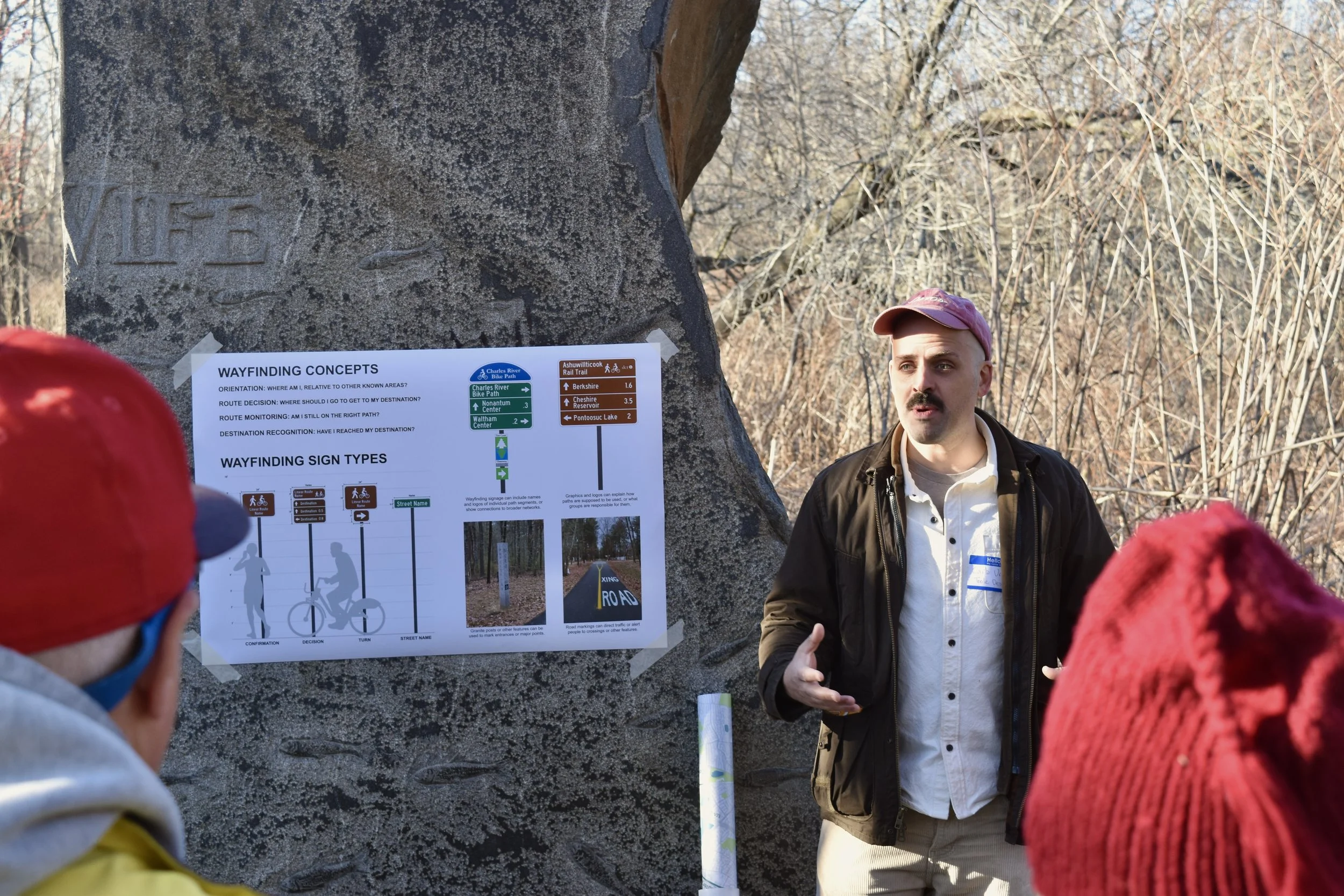A person standing outdoors near a large rock presenting a graphic titled "Wayfinding Concepts" to a group of people in the foreground