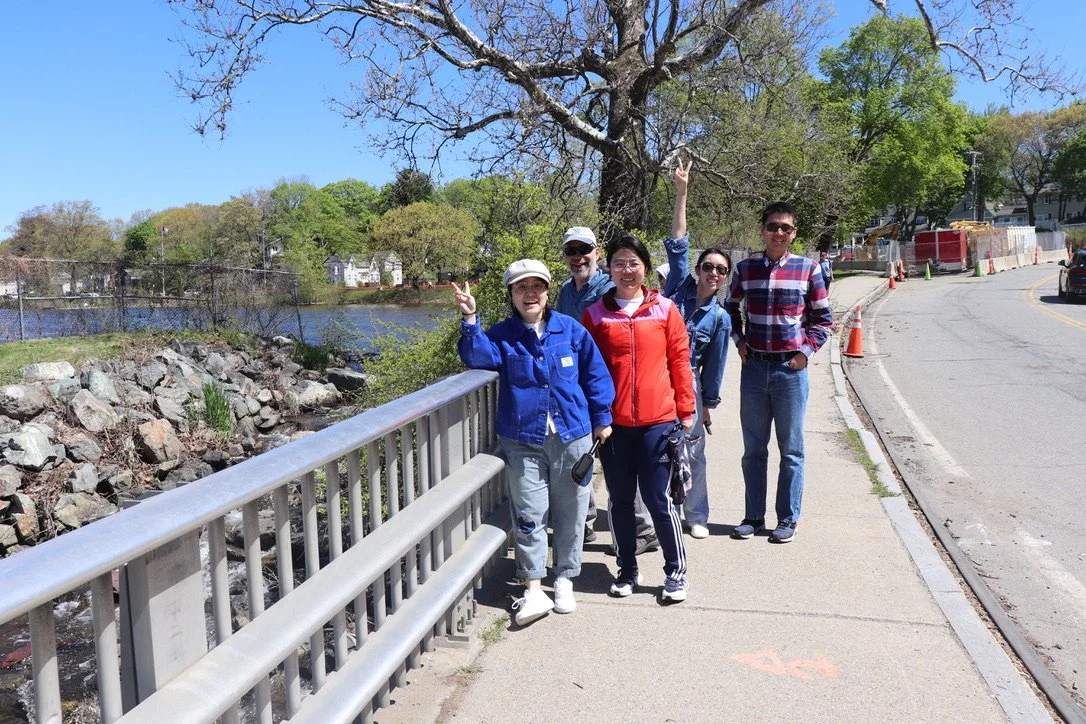 A group of five people stands by a fence on the side of a road smiling and making peace signs to the camera. Over their right shoulder you can see Woburn's Horn Pond and the Scalley Dam spillway, where river herring gather