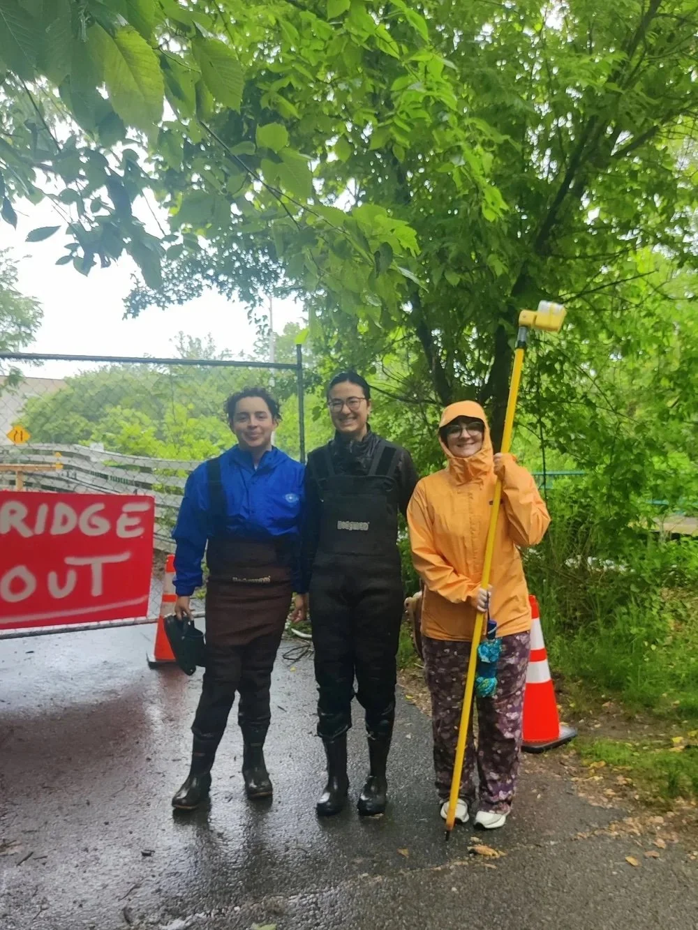Juan, Kenna, and Ry pose for a photo. They are dressed for rain and are holding a long yellow sampling pole.