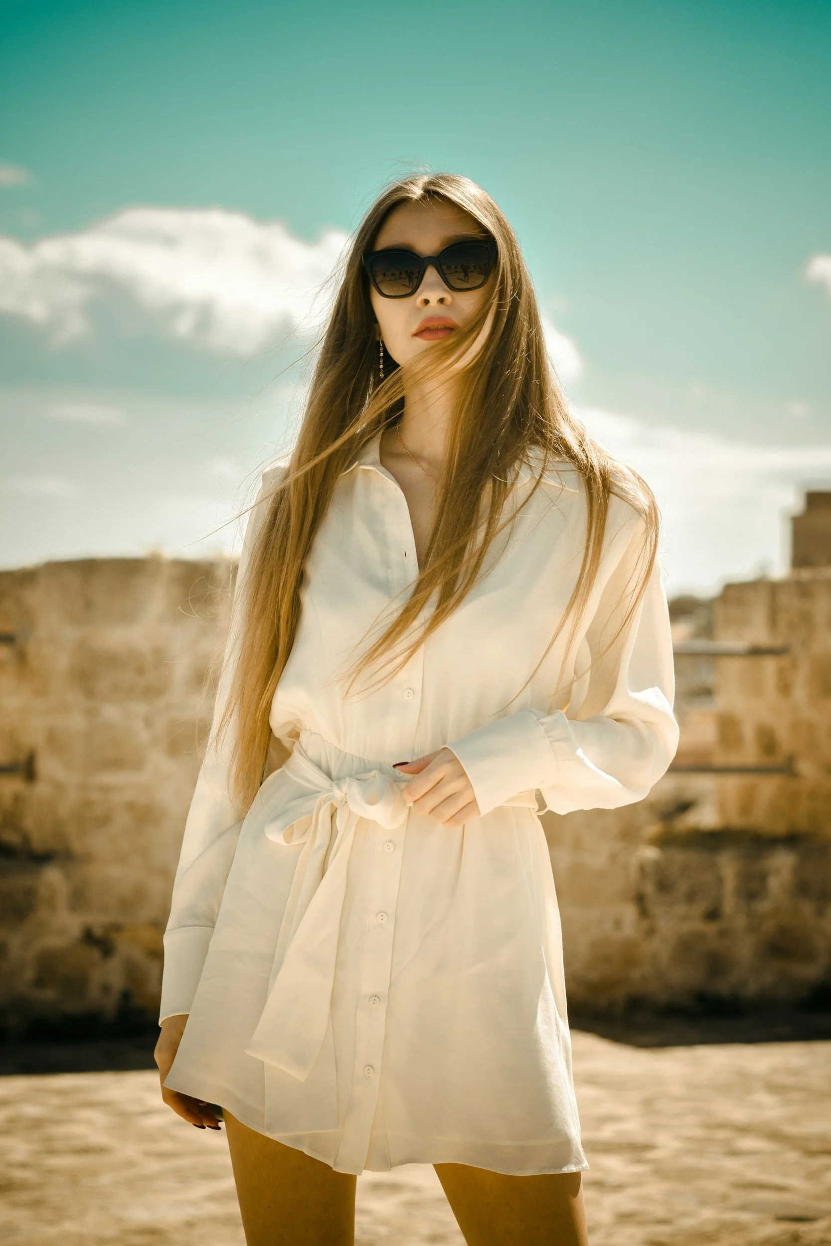 Young woman wearing a white shirt dress and sunglasses, standing outdoors with ruins and a partly cloudy sky in the background.