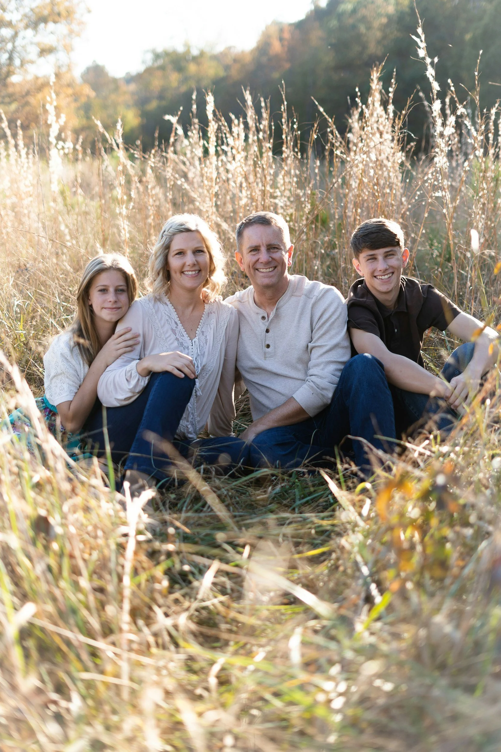 Family of four sitting in a grassy field during autumn, smiling at the camera. The family includes a mother, father, daughter, and son, surrounded by tall, dry grass and trees in the background.
